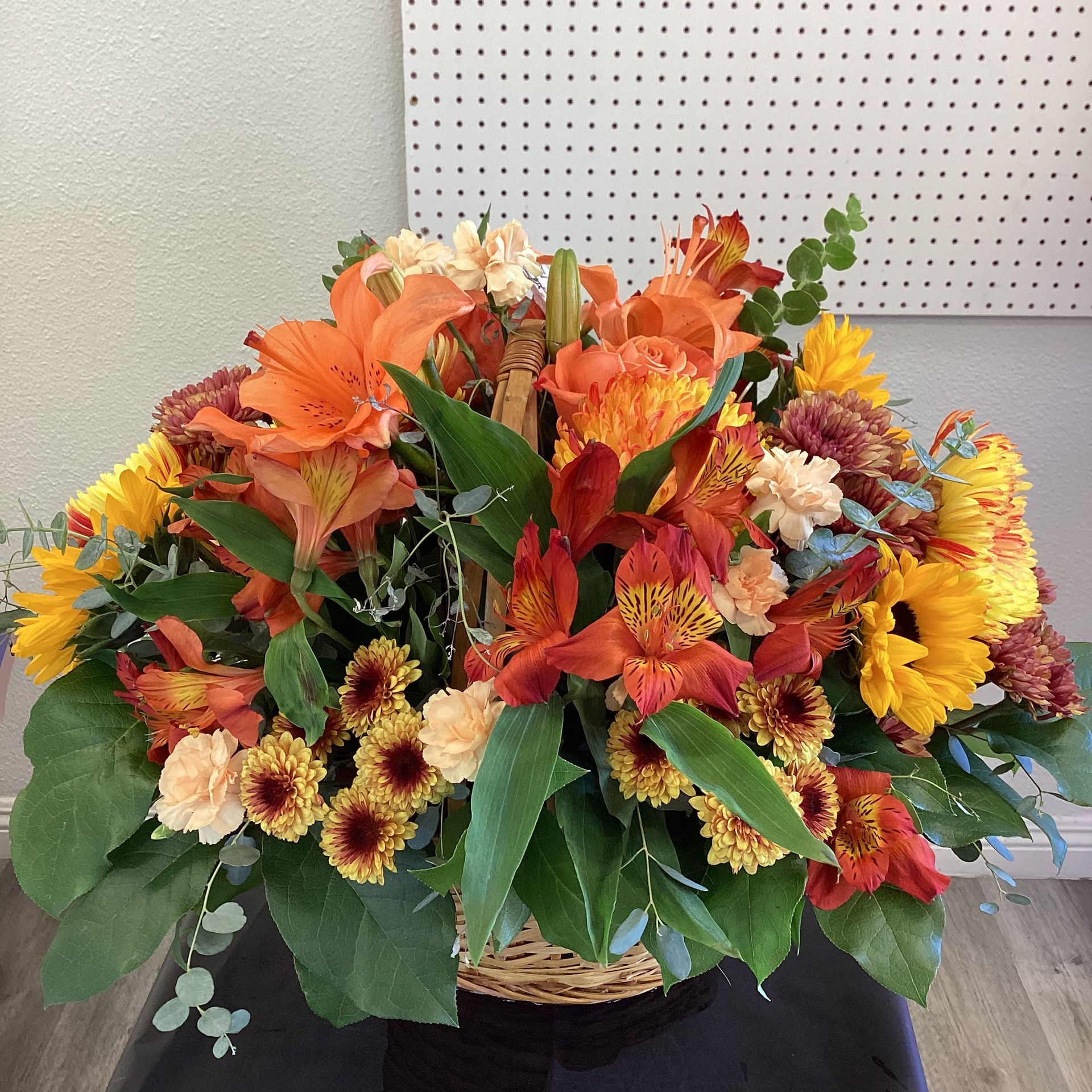 A vibrant floral arrangement in a basket featuring orange lilies, sunflowers, yellow mums, and greenery against a wall.