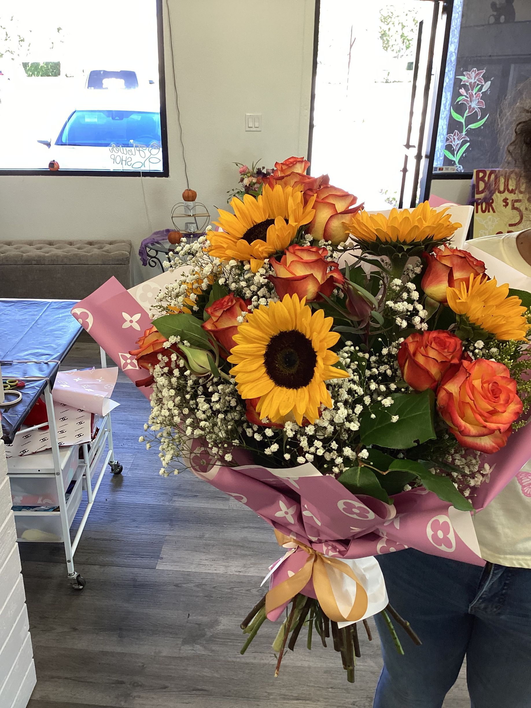 A bouquet of sunflowers and orange-tipped roses wrapped in pink floral paper held by a person.