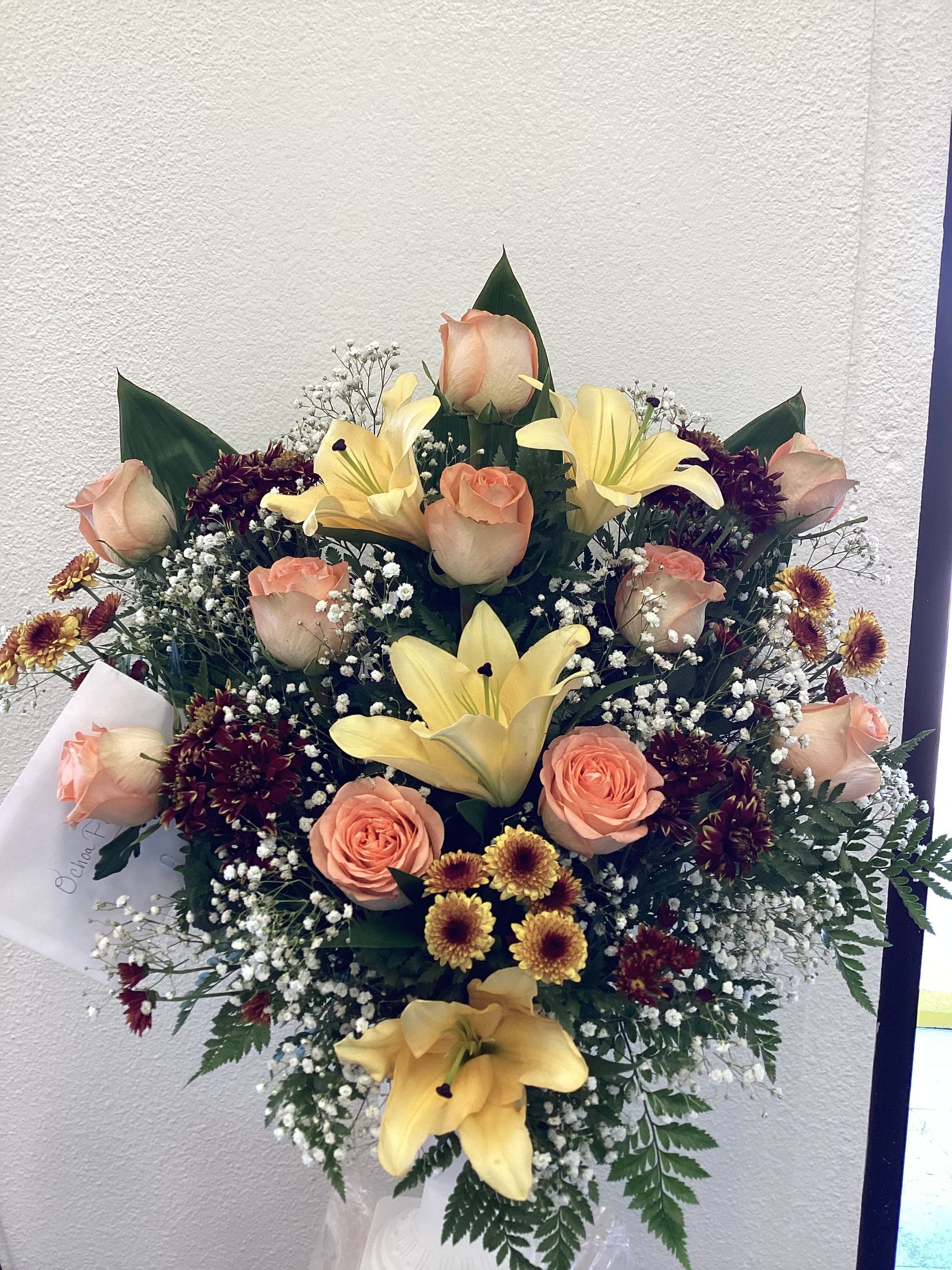 A bouquet of peach roses, yellow lilies, and dark red carnations with white baby’s breath and greenery.