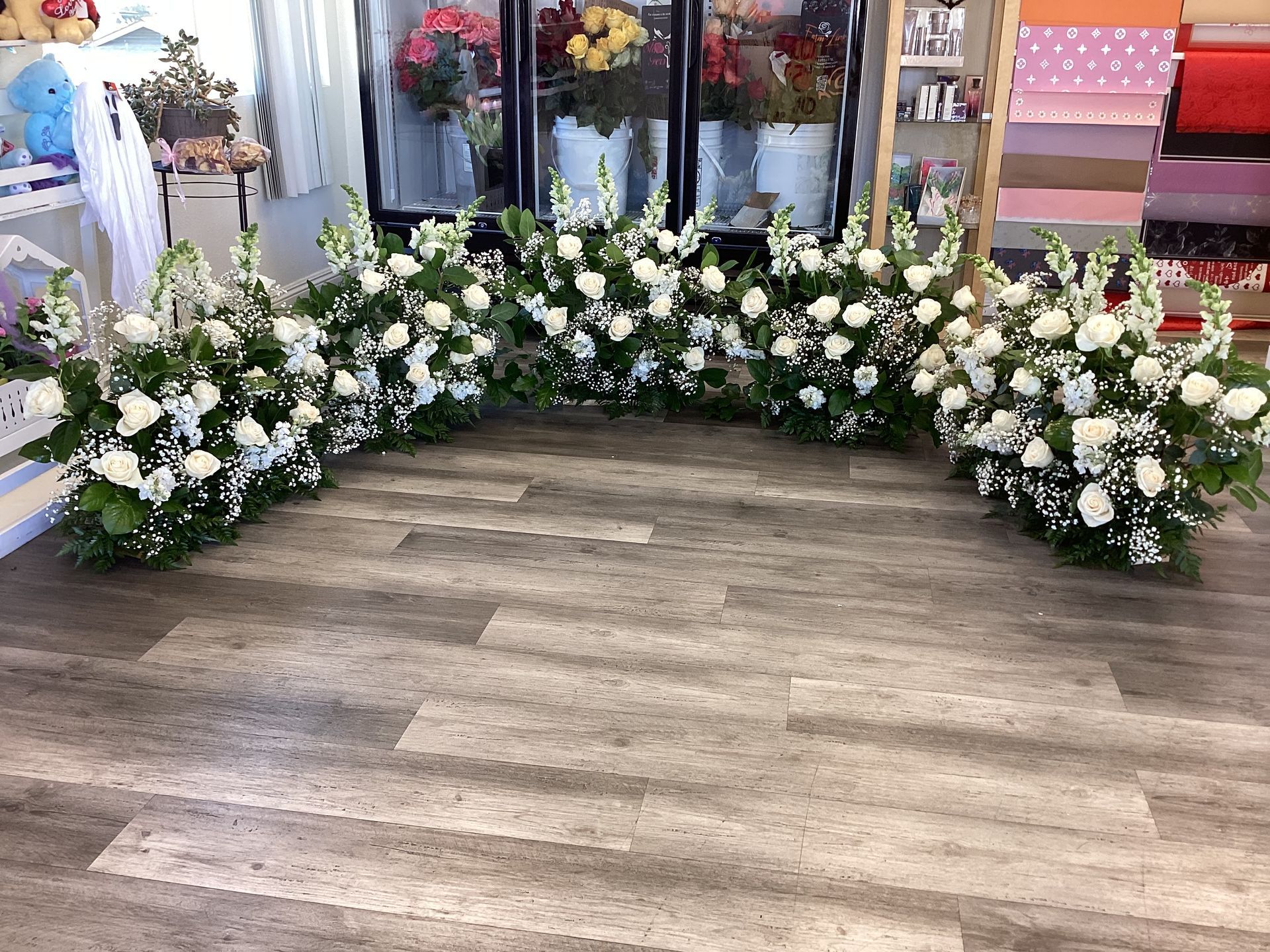 Five large, lush floral arrangements composed of white roses and tall snapdragons, arranged in a wide arc on a wood floor.