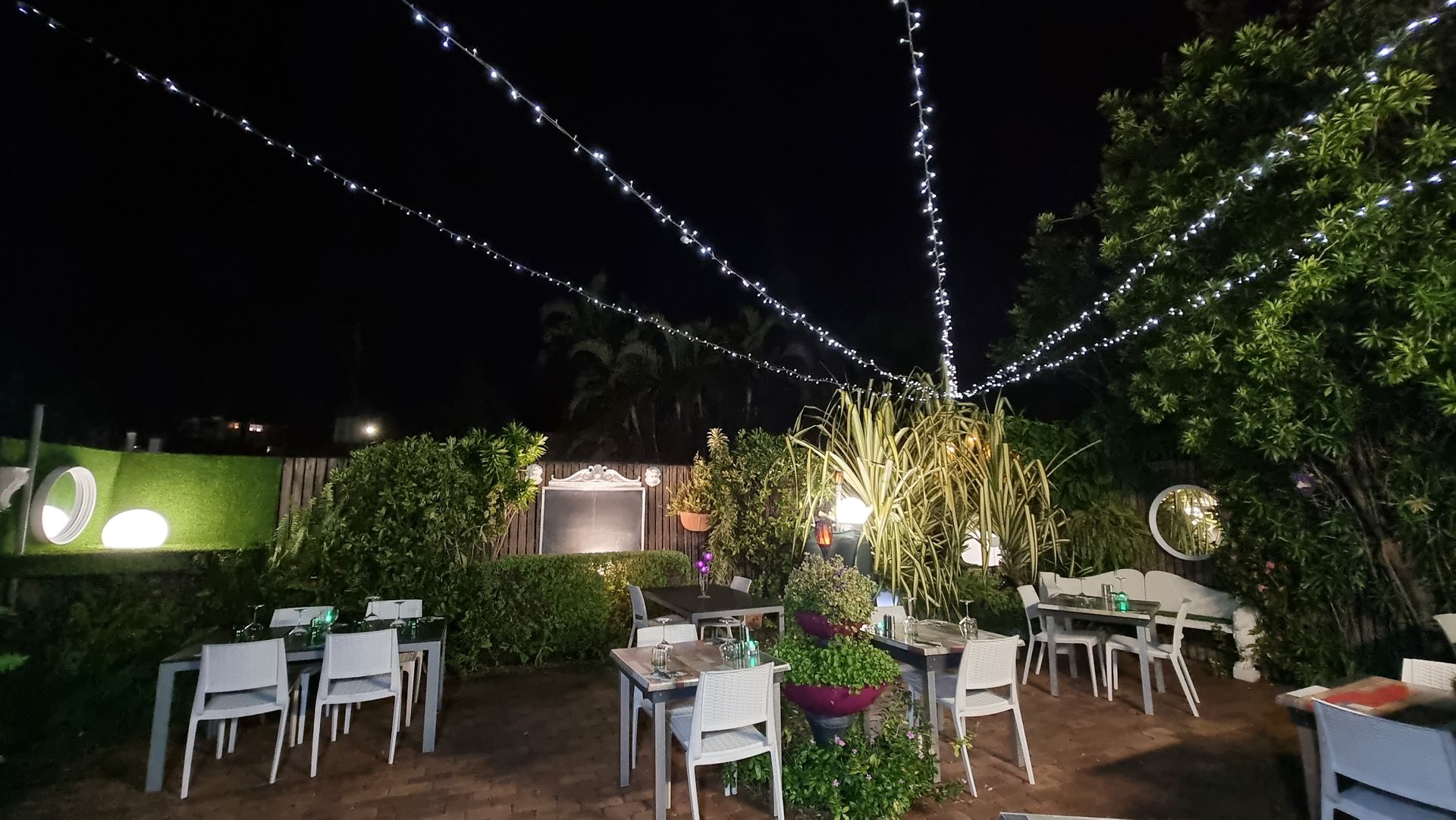 A patio with tables and chairs at night with lights hanging from the ceiling  — Wayne & Adele's Garden of Eating BYO Restaurant In South Townsville, QLD