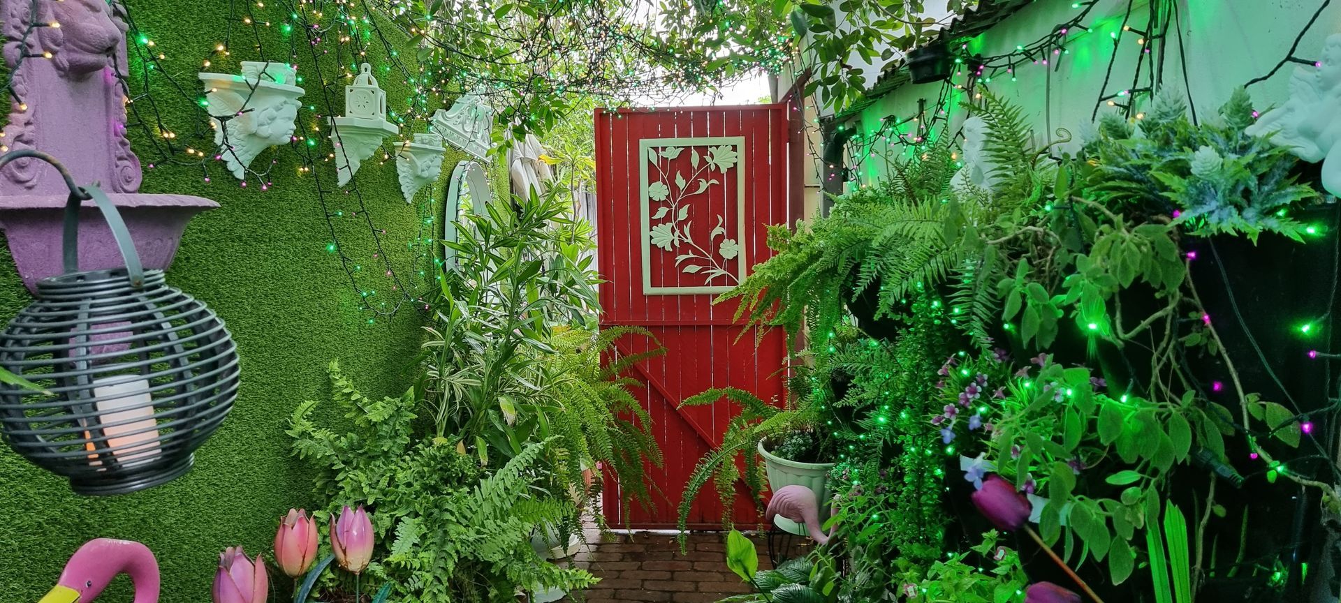 A red door is surrounded by lots of green plants and flowers  — Wayne & Adele's Garden of Eating BYO Restaurant In South Townsville, QLD