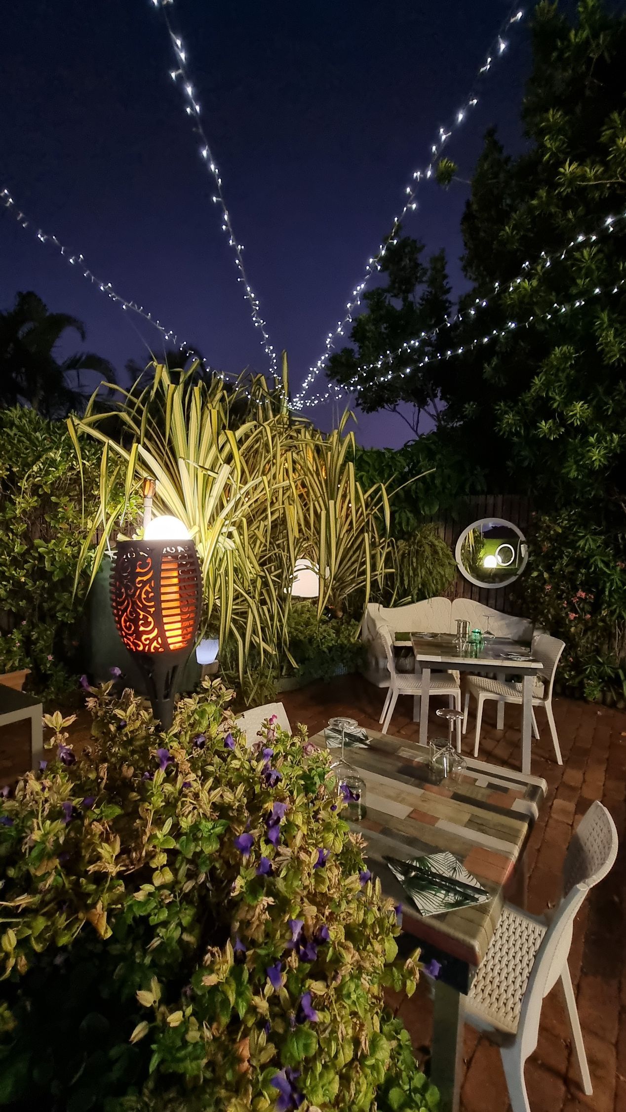 A patio with a table and chairs at night with lights hanging from the trees  — Wayne & Adele's Garden of Eating BYO Restaurant In South Townsville, QLD