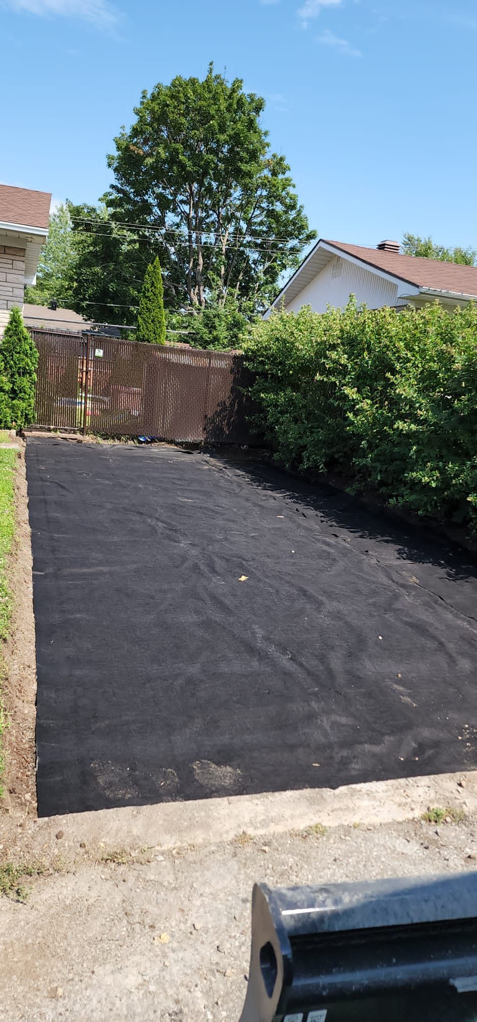 Une allée en asphalte noir avec une clôture en arrière-plan et une maison en arrière-plan.