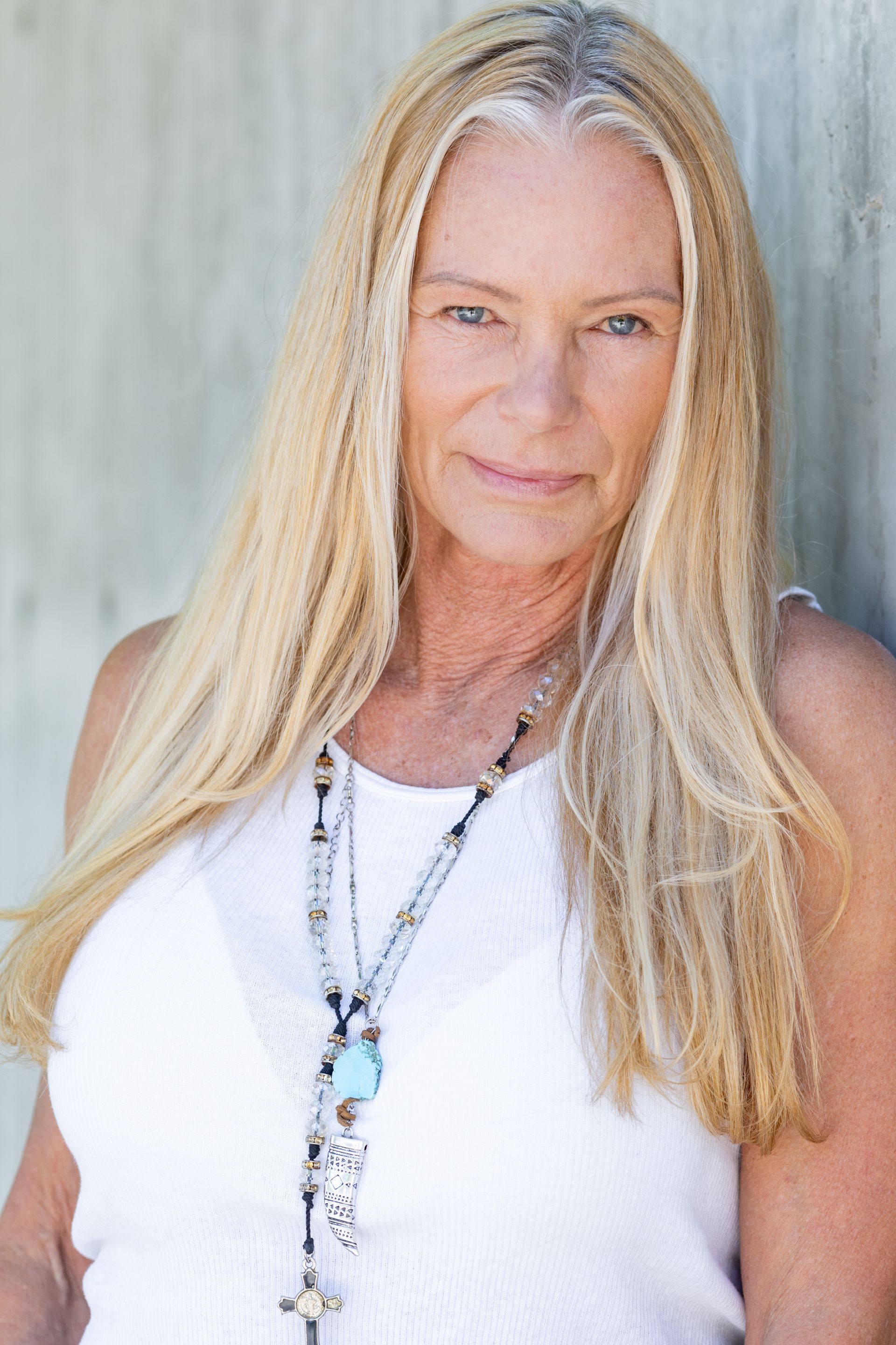 A woman with long blonde hair is wearing a white tank top and a necklace.