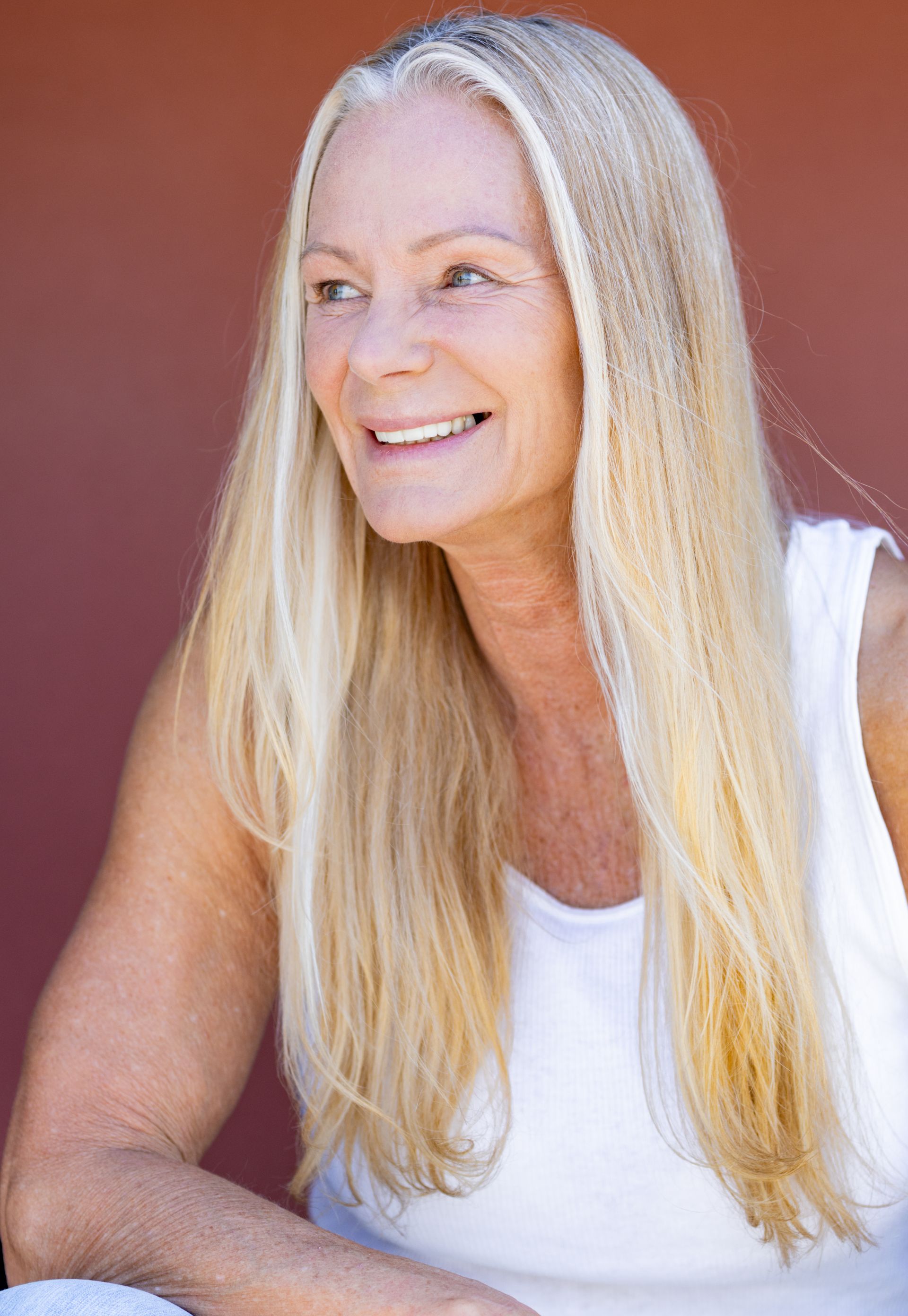 A woman with long blonde hair is wearing a white tank top and smiling.