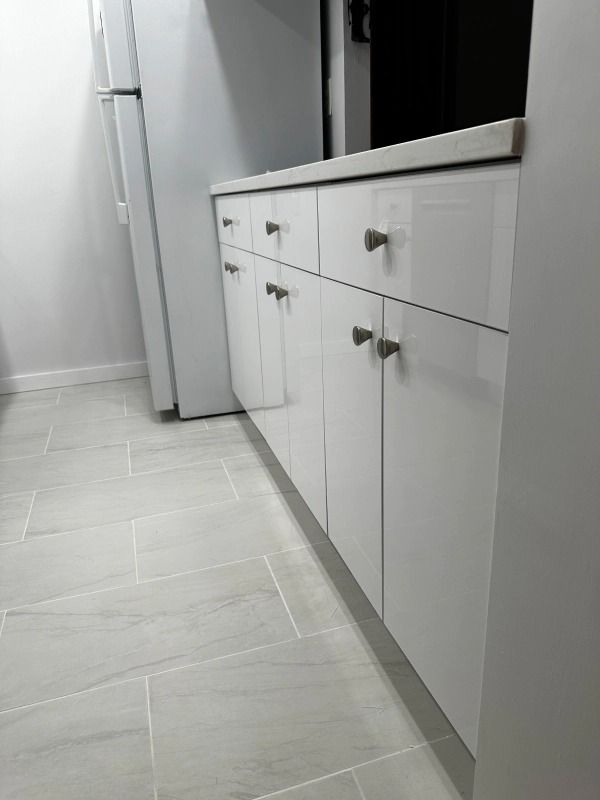 White kitchen cabinets with silver hardware and a white countertop, next to a white refrigerator.