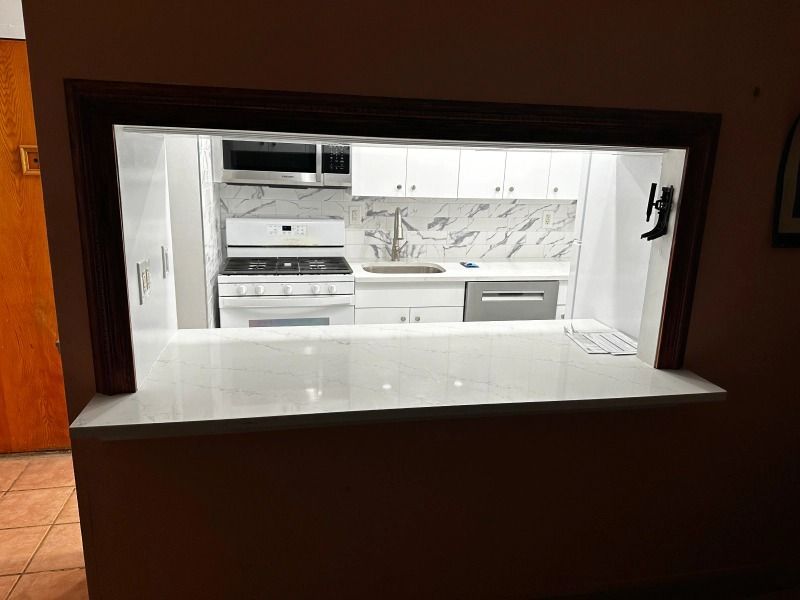 White kitchen seen through a pass-through, with white counters, cabinets, appliances, and marbled backsplash.