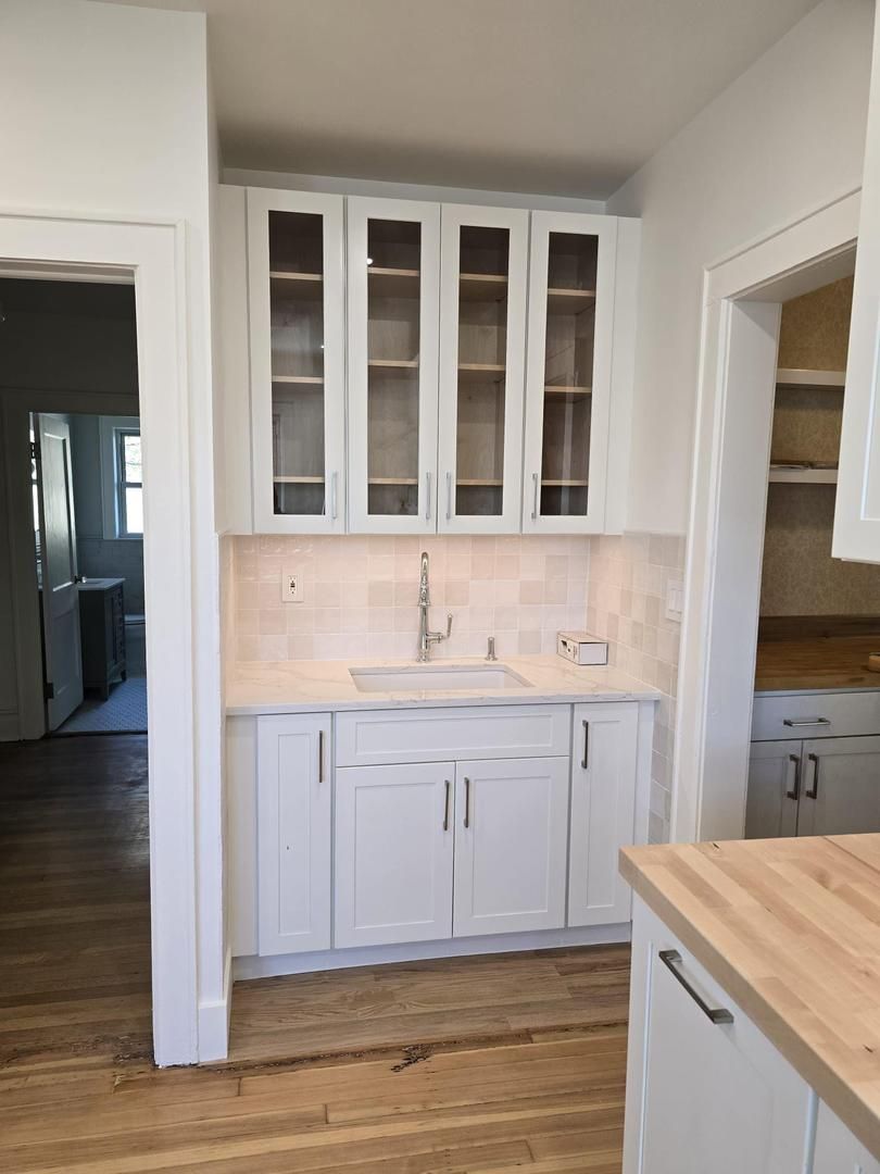 White kitchen cabinetry with glass-paned uppers and sink, wood floor, doorway in the background.