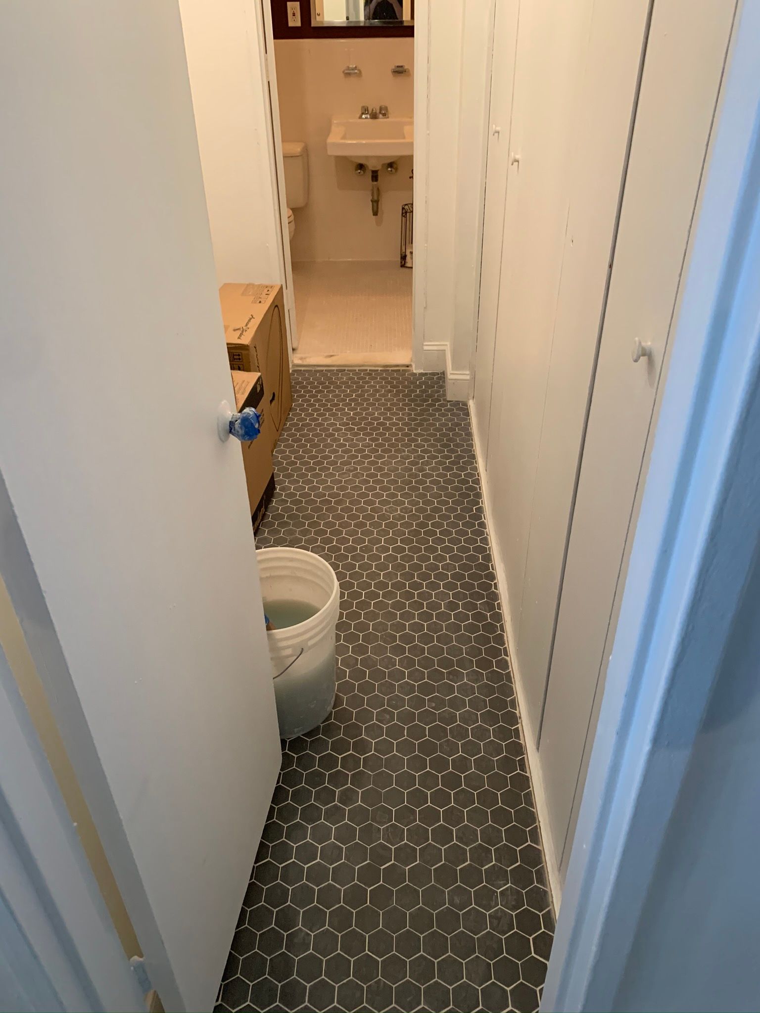 Narrow hallway with dark hexagon tile floor, leading to a bathroom. A bucket and cabinet are visible.