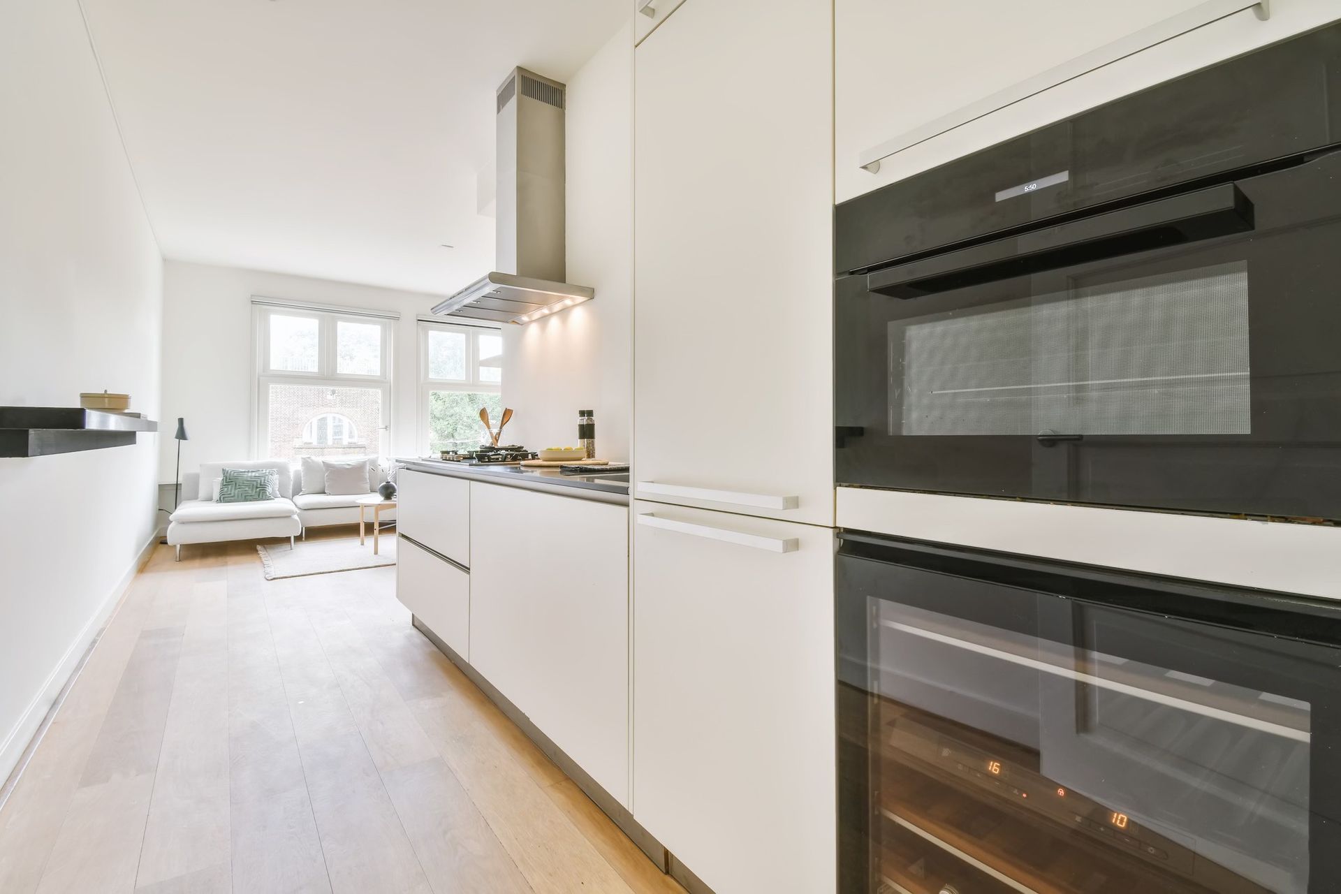Modern white kitchen with built-in appliances, light wood floor, and view into a living room.