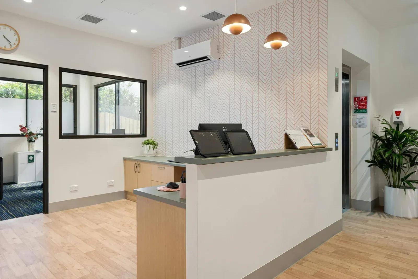 Reception area with white desk, two monitors, wood floor, patterned wall, and potted plant.