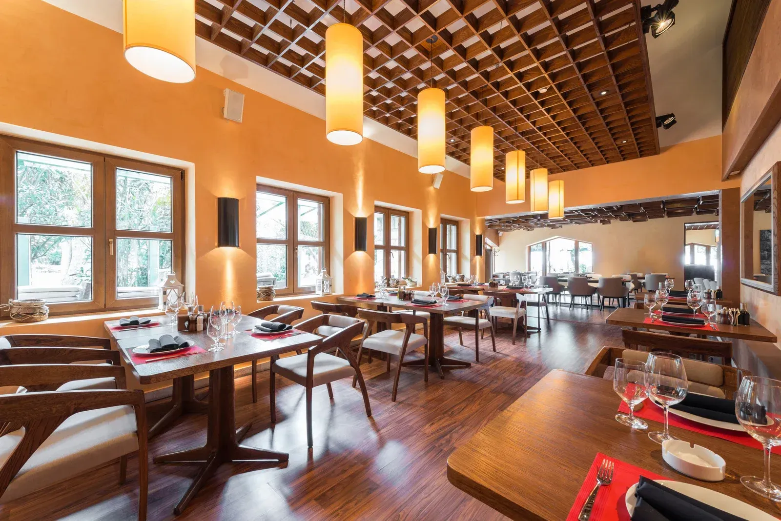 Restaurant interior with tables, chairs, windows, and hanging lights. Orange walls, wooden floor, and grid ceiling.