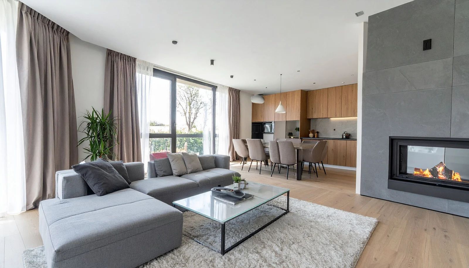 Modern living room with grey sectional sofa, fireplace, and open kitchen with wood cabinets.