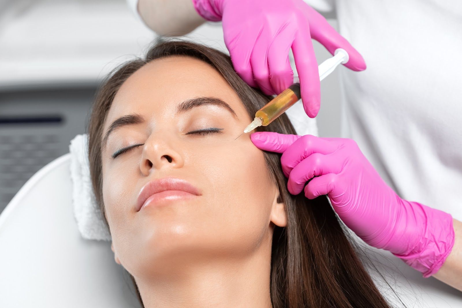 Woman receiving facial injection near eye; hands in pink gloves holding syringe.