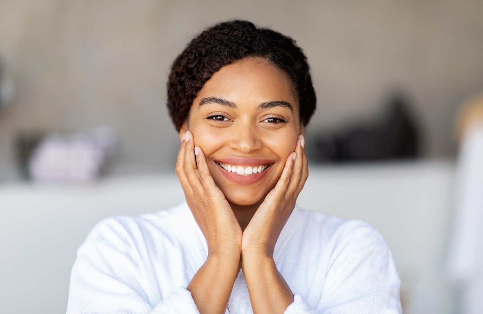 Woman in white robe smiles, holding her cheeks in a bathroom setting.