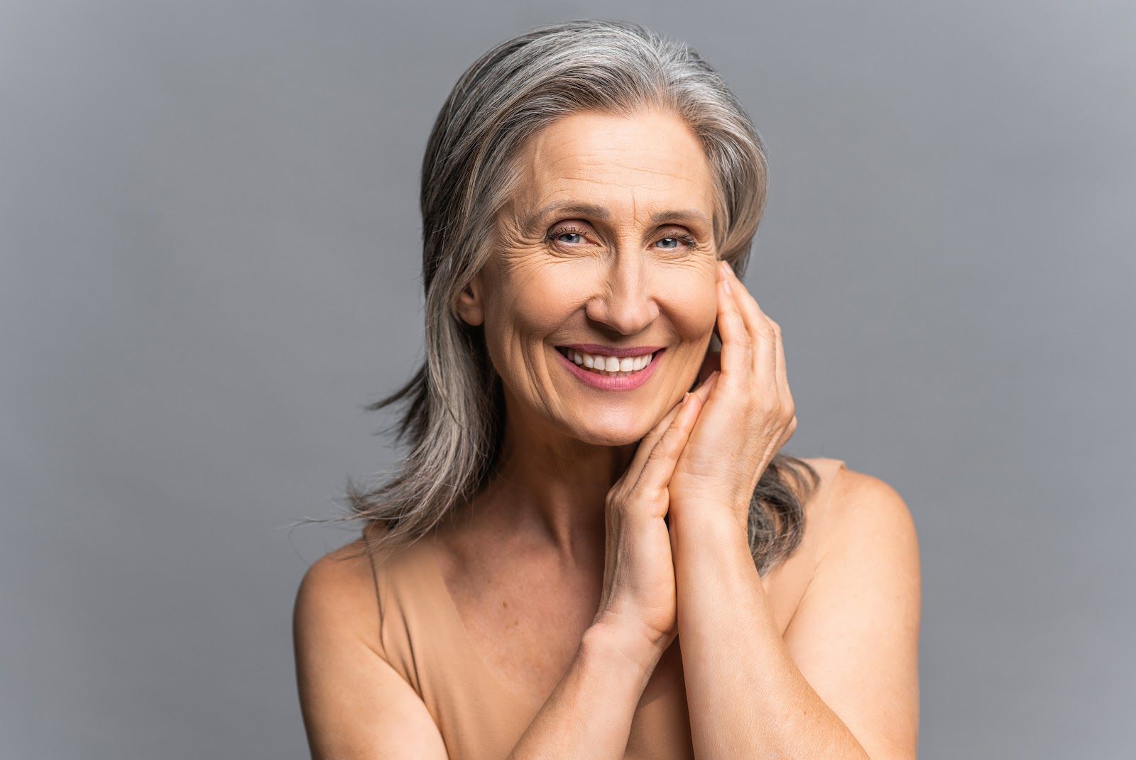 Woman with gray hair smiles, touching her face with both hands, neutral background.