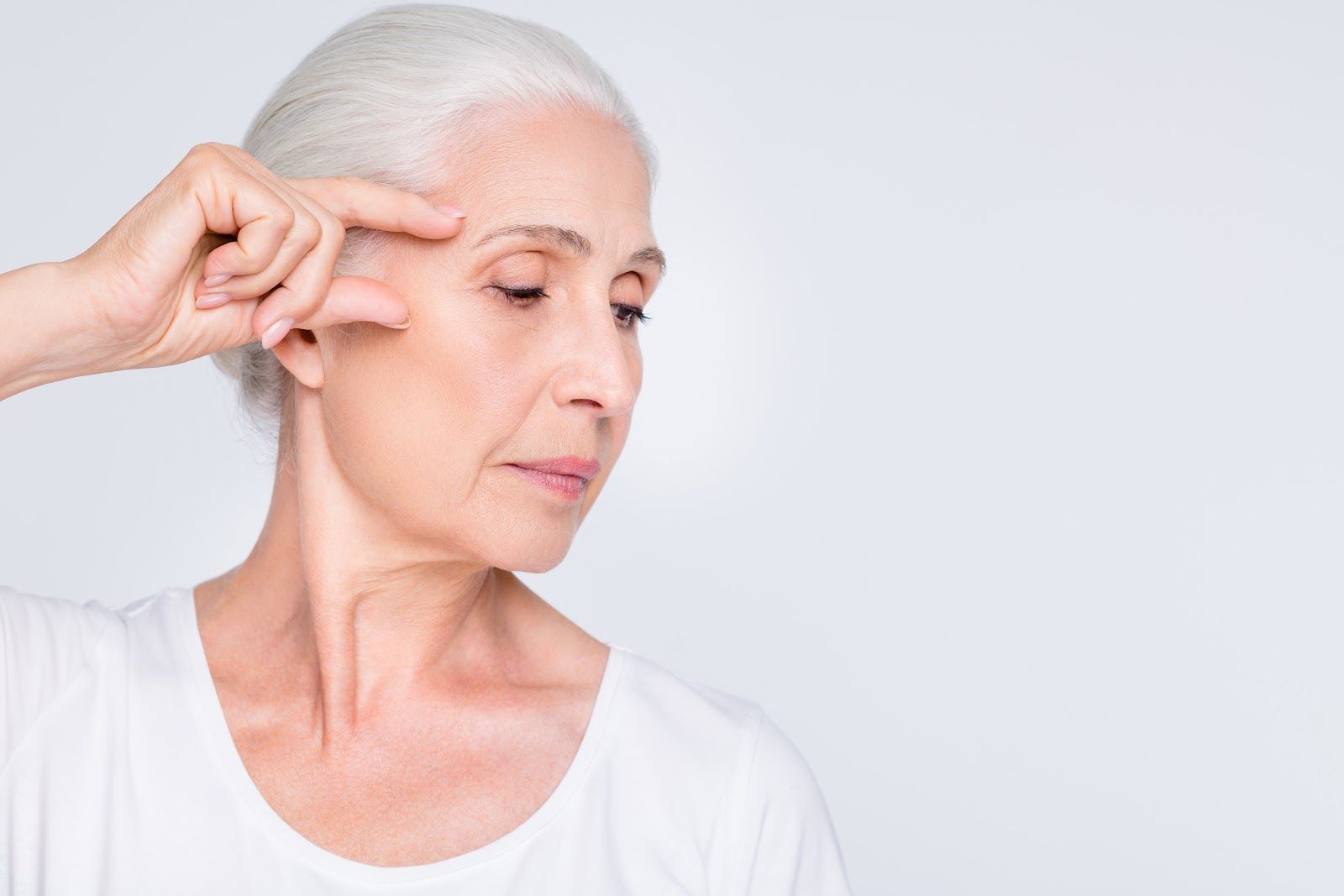 Woman in white top, touching her face, looking downward with a neutral expression.