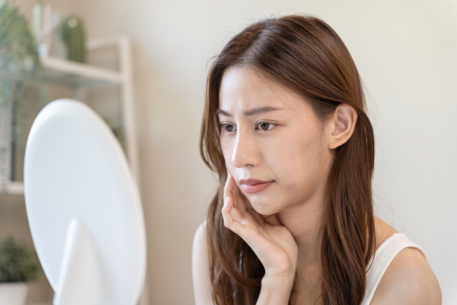 Woman looking in a mirror, touching her cheek with concern. Light skin, brown hair, white top, bright room.