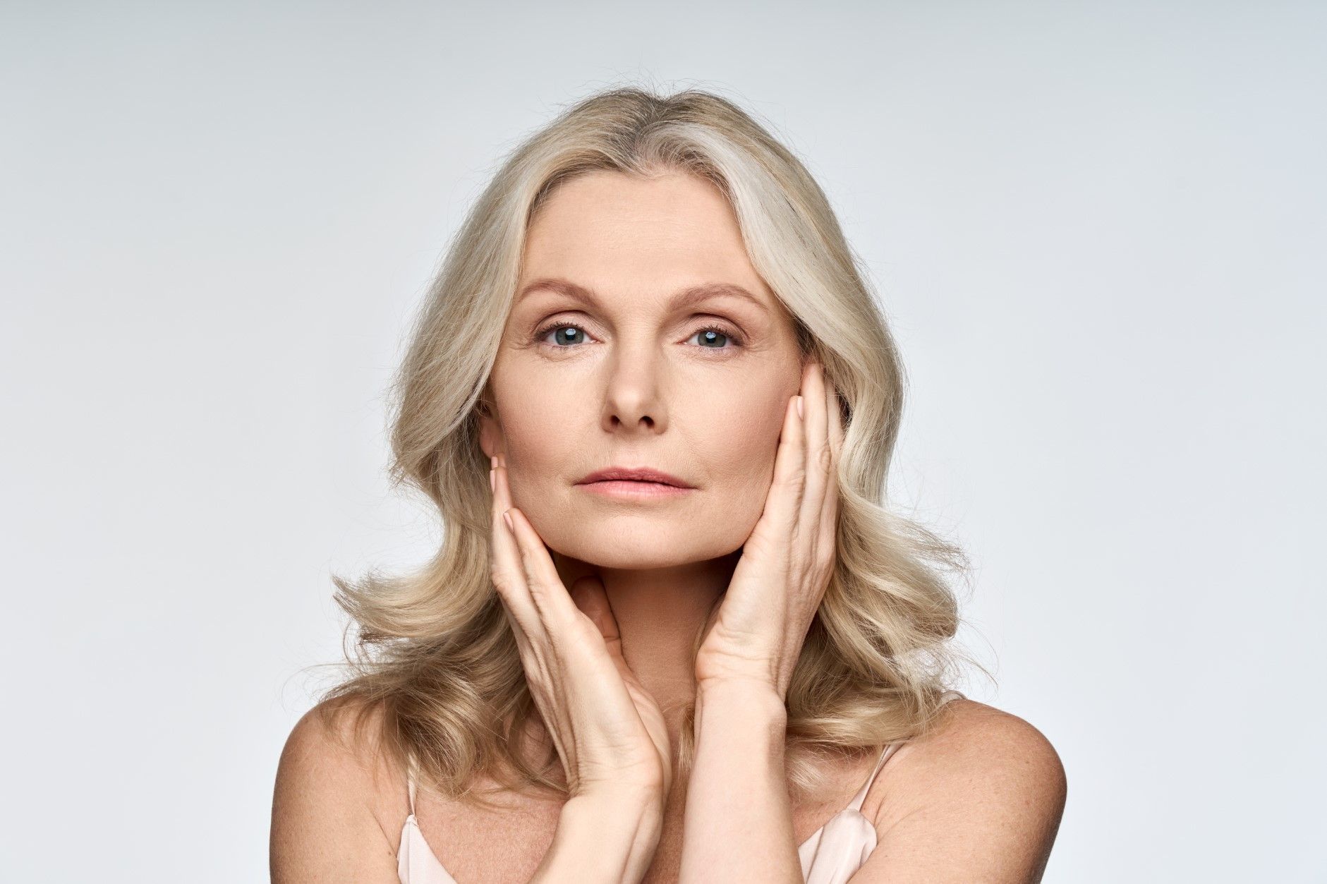 Woman with fair skin, touching her face with both hands, light background.