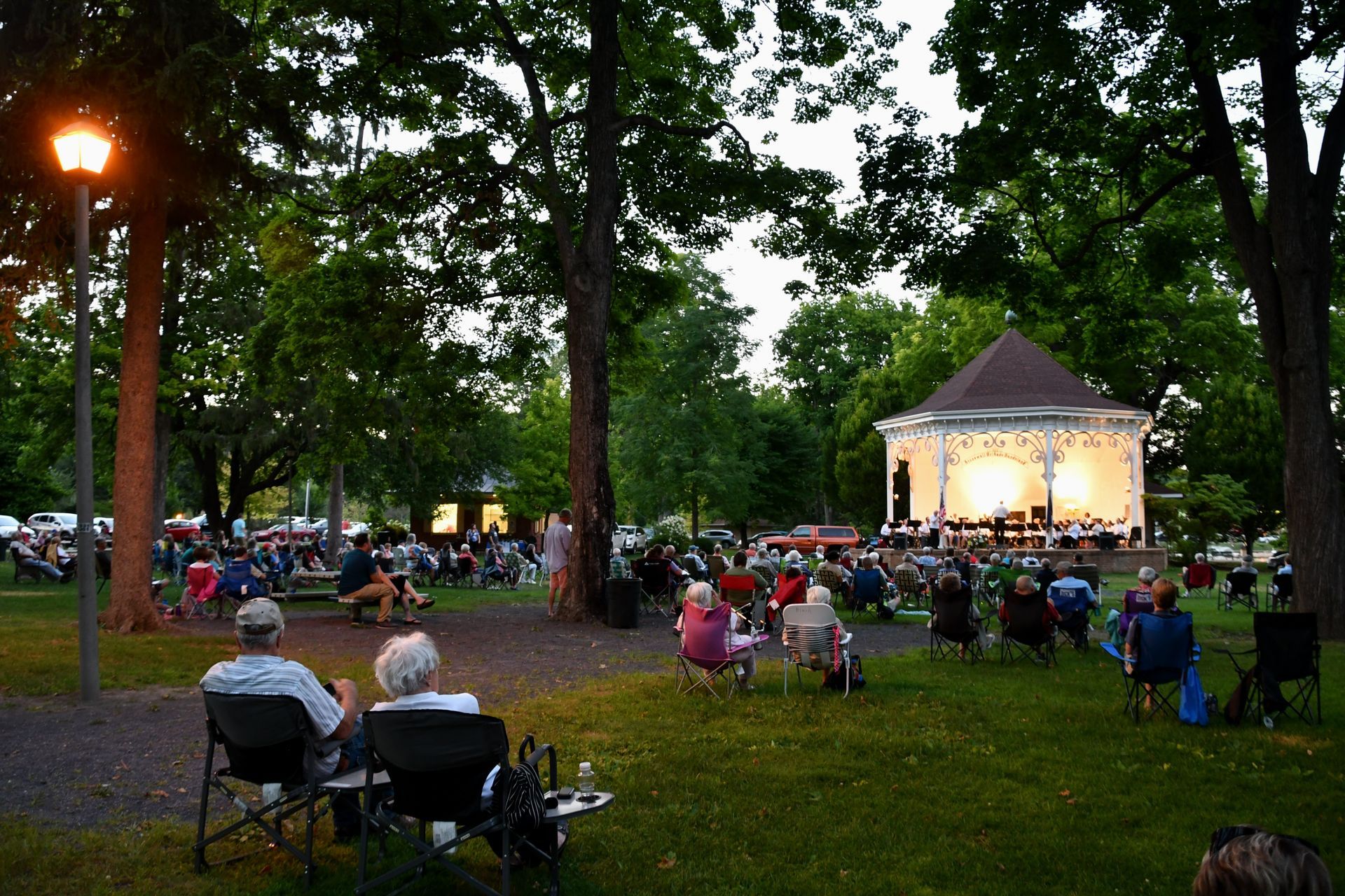 People seated in park, watching a concert at a lit gazebo at dusk.