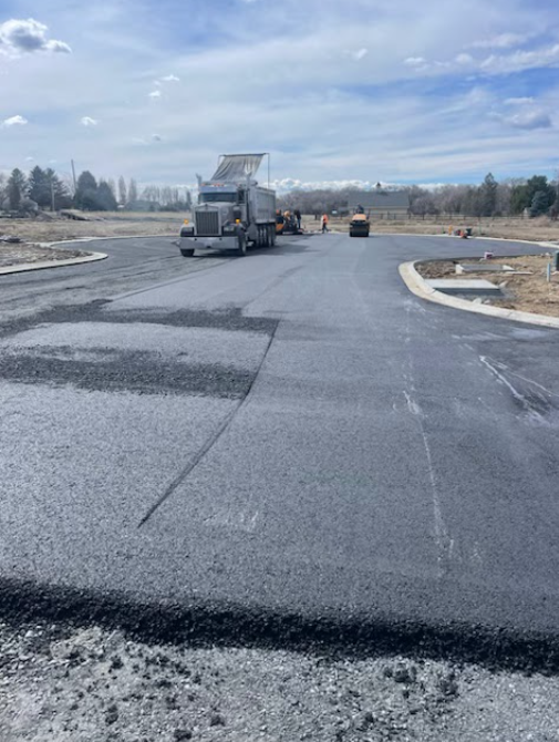 A construction site with a dump truck and a steamroller working on a freshly paved asphalt road.