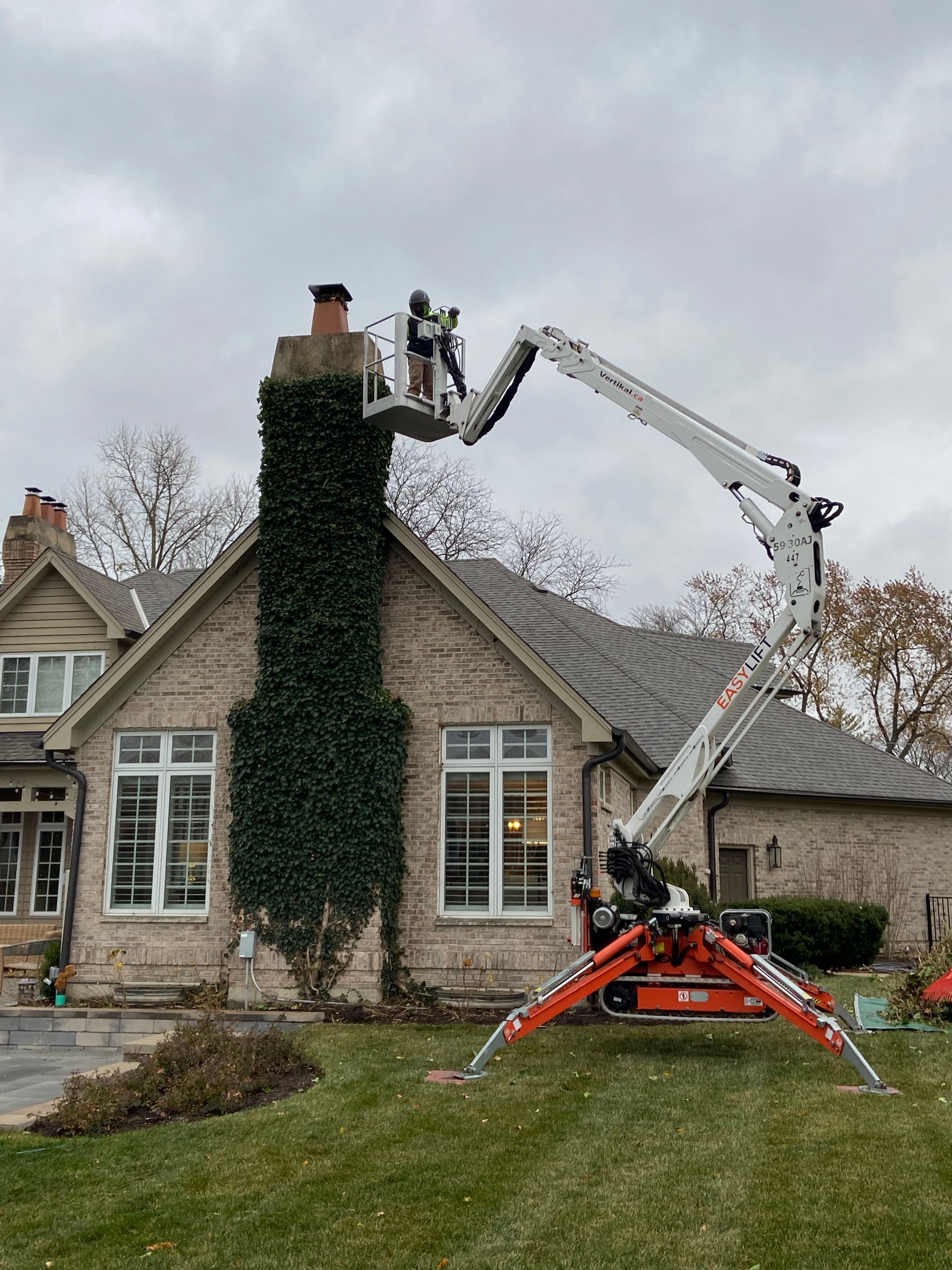 A worker in a lift basket is inspecting a chimney covered in ivy, located next to a house.