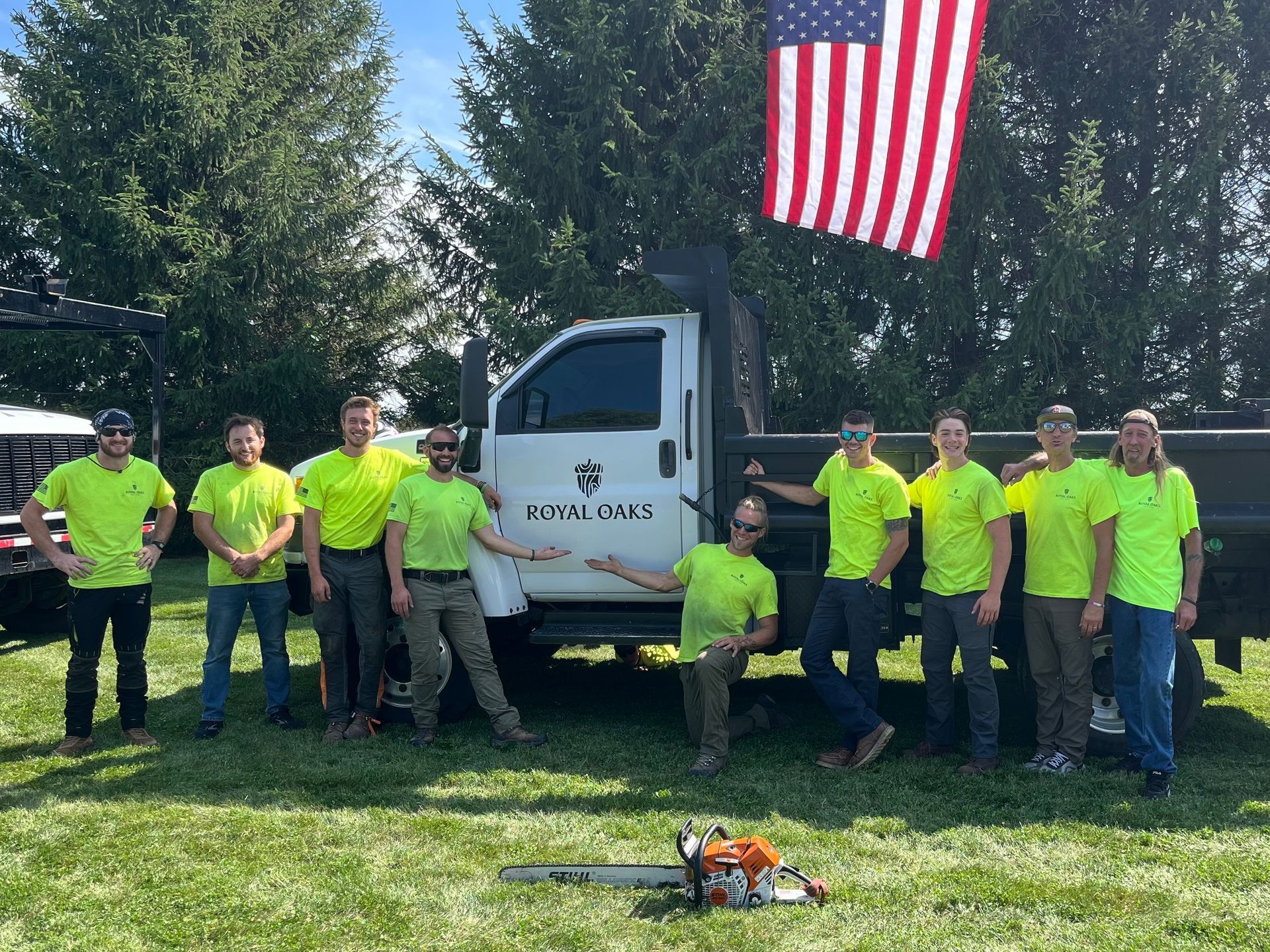 A team of tree service workers in lime green shirts poses with a truck and American flag on a lawn.
