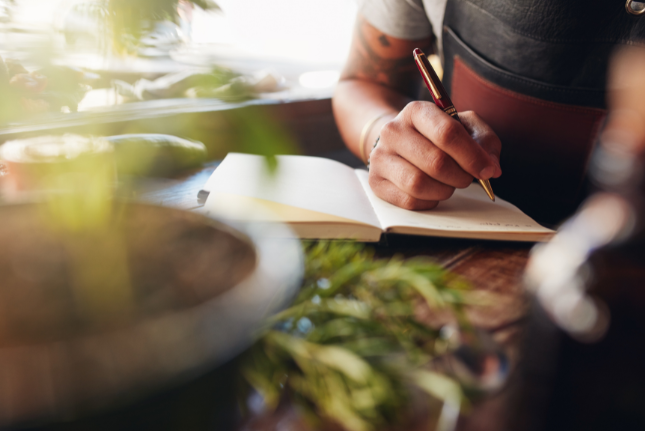 Person with tattoos writing in a notebook at a wooden table. Herbs in the foreground.