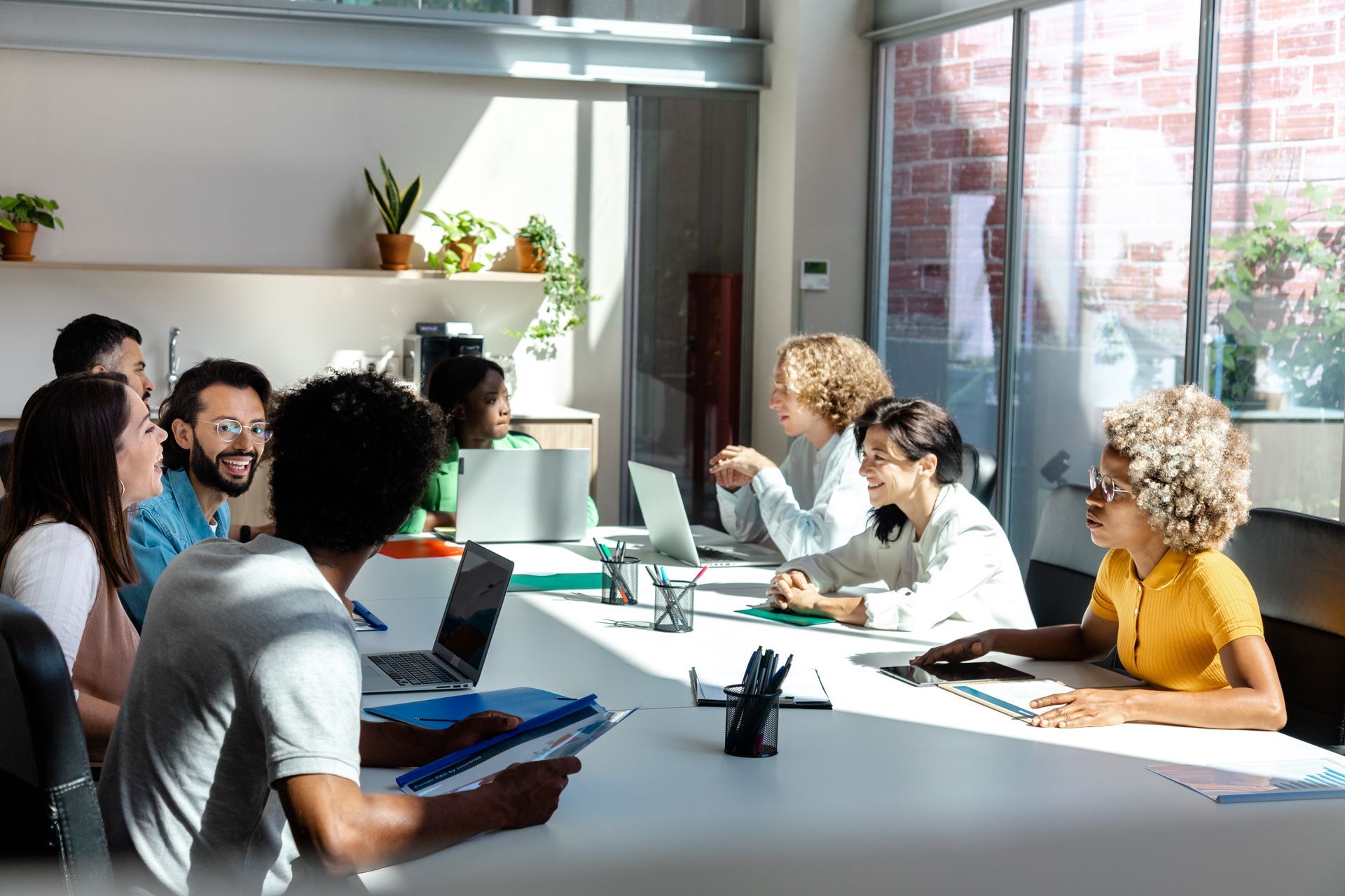 People in a bright office meeting at a long table, working on laptops and papers.