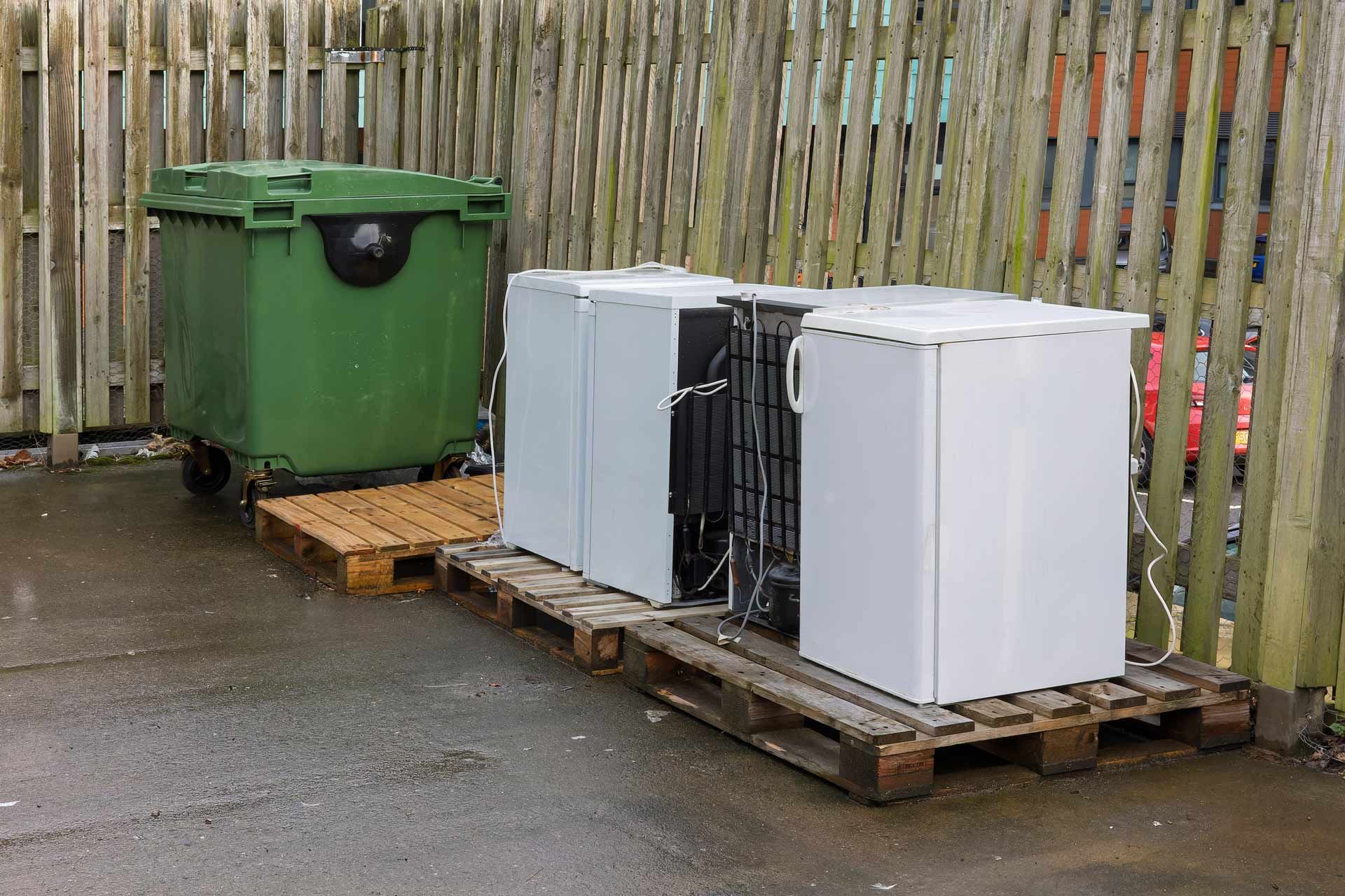 Green trash bin and white refrigerators on pallets against a wooden fence.