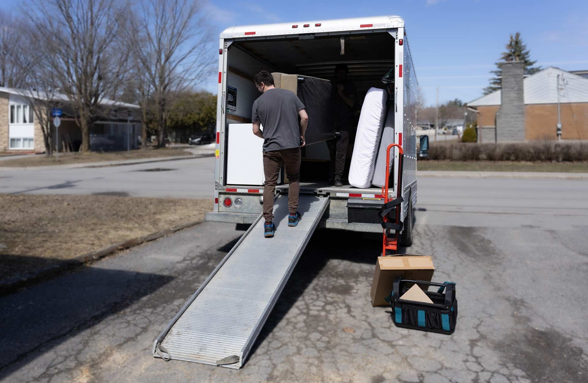 Man walks up ramp into moving truck, boxes and mattress visible, outside on a sunny day.