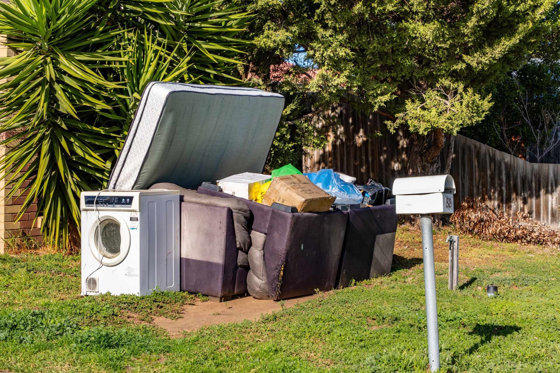 Pile of household waste, including a mattress and washing machine, overflowing a waste bin outdoors, next to a mailbox.