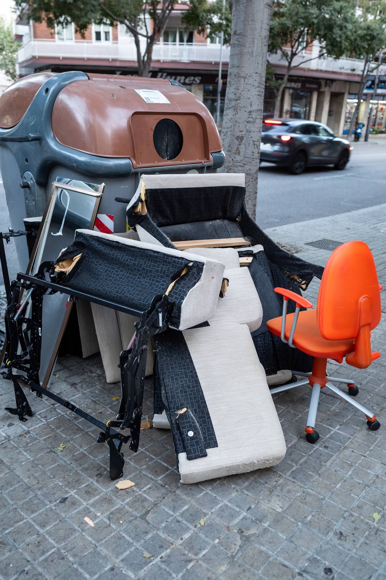 Dumpster with discarded furniture including an office chair, and pieces of a sofa. Street scene.