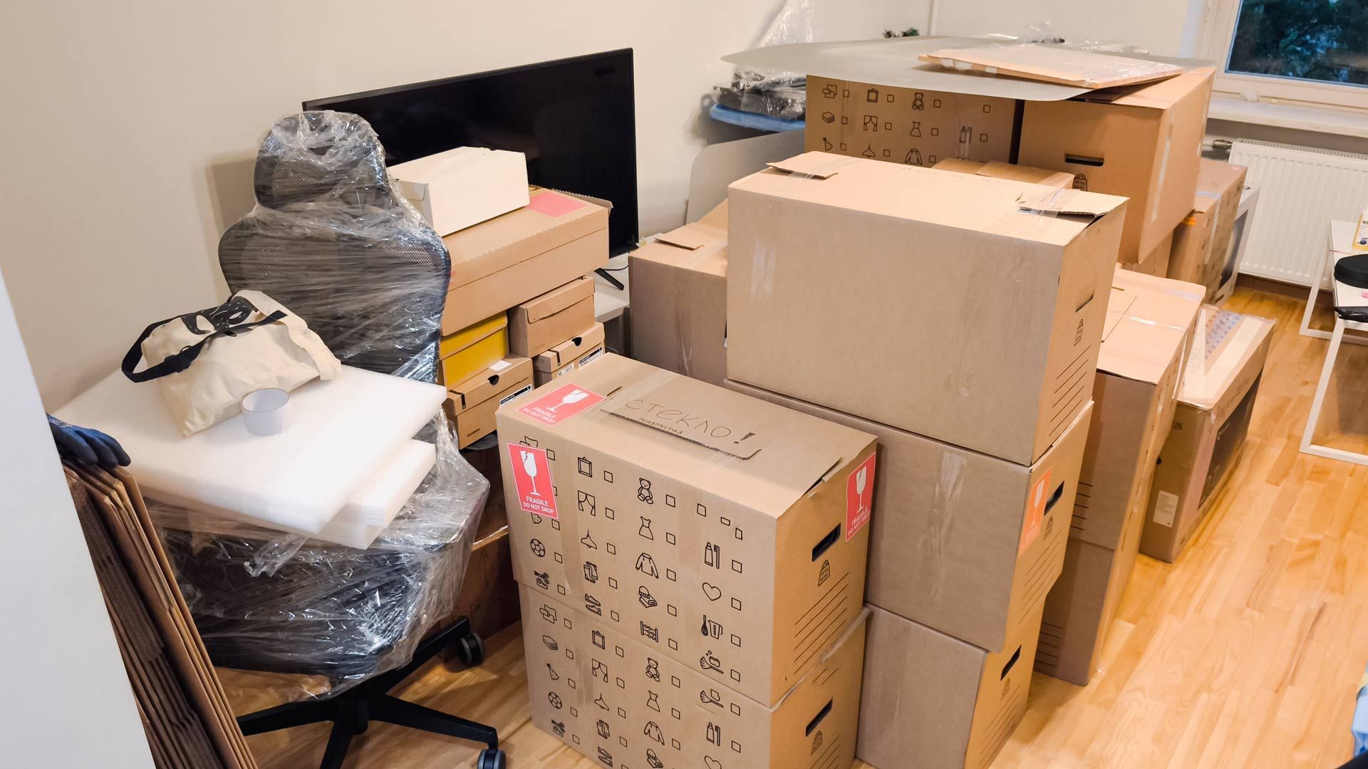 Cardboard boxes stacked in a room, ready for moving. A wrapped office chair sits nearby.
