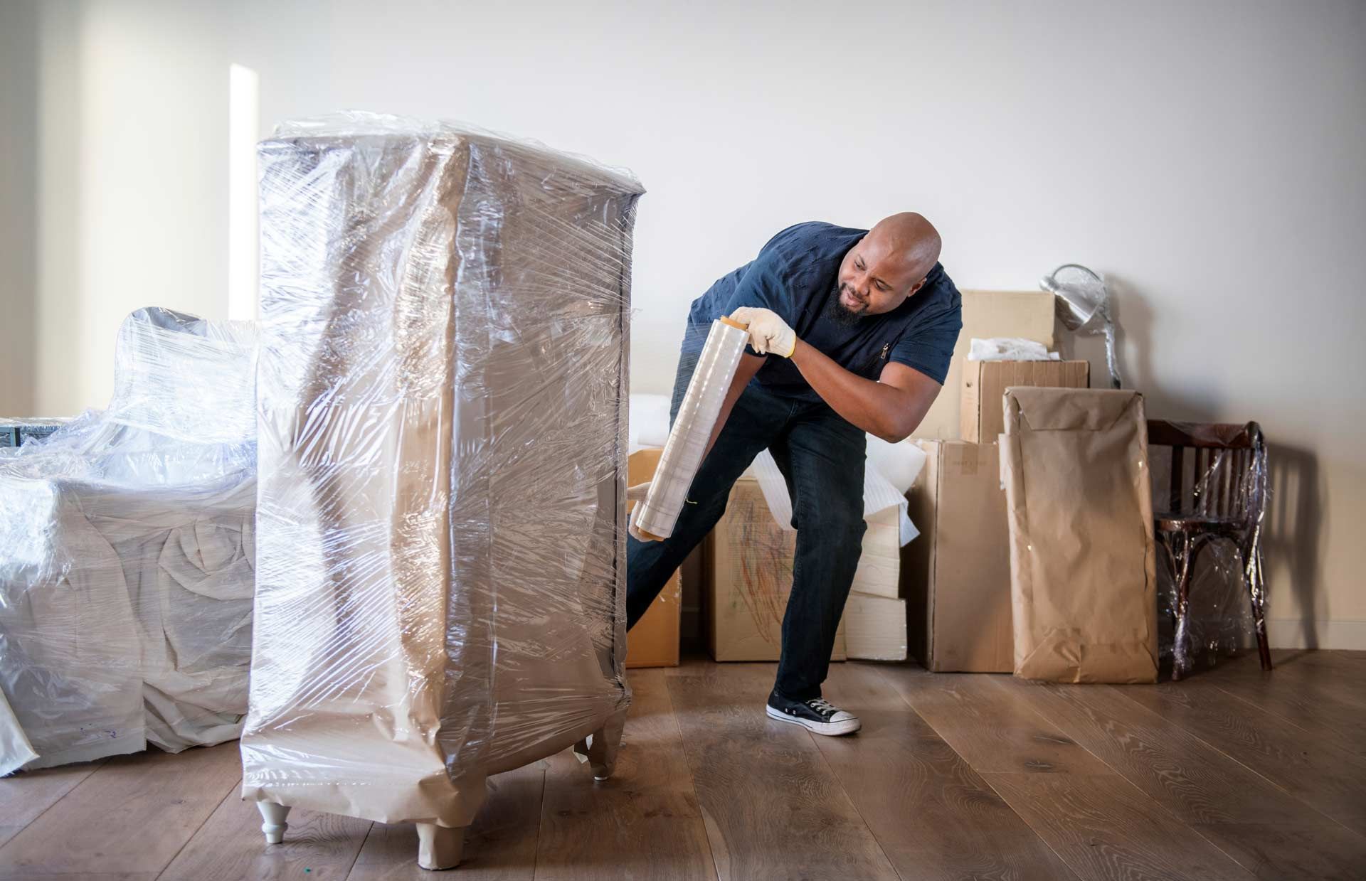 Man wrapping furniture in plastic, preparing for a move. Inside a room with wrapped items and wooden floor.