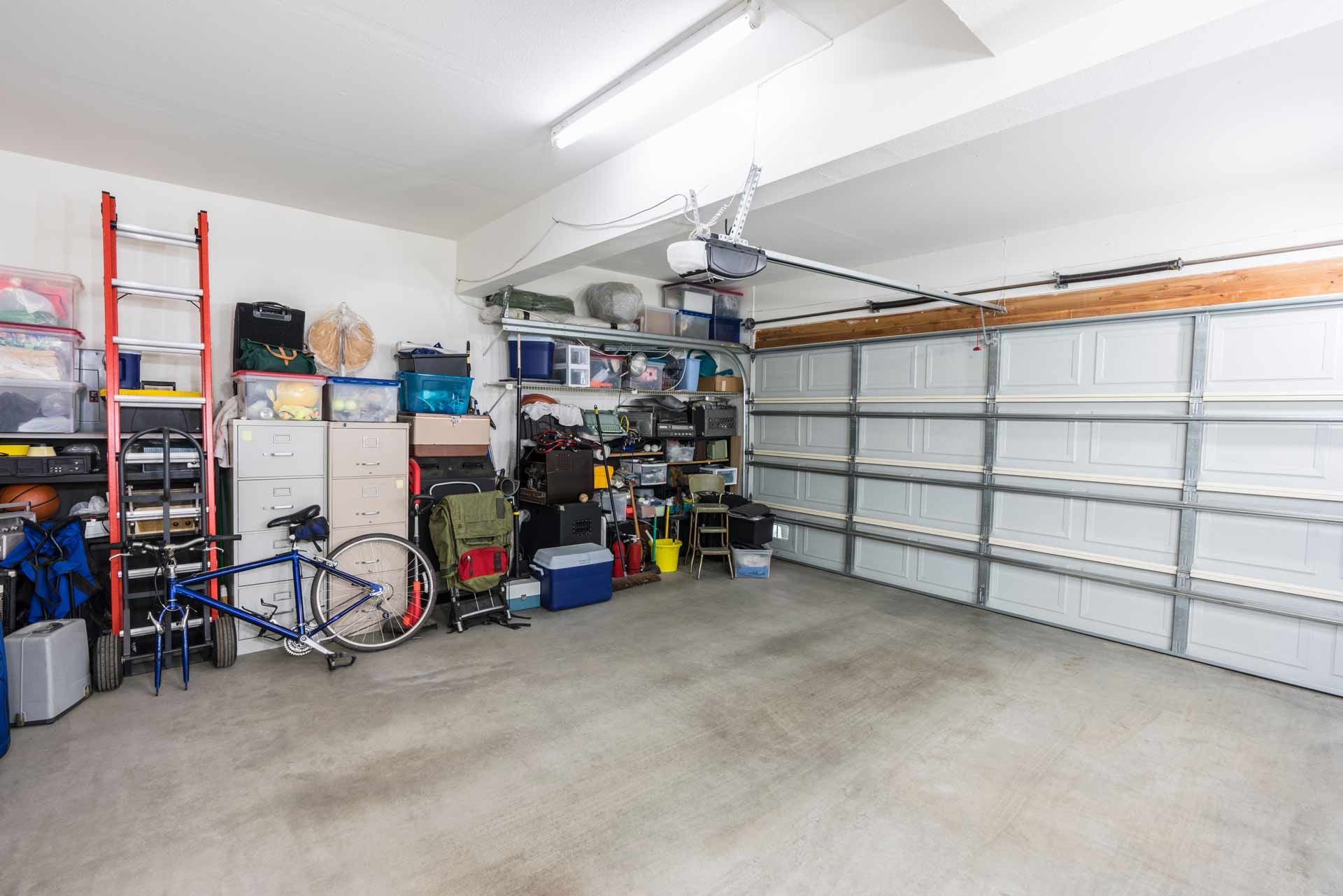 Cluttered garage interior with storage shelves, a bicycle, and various items. Bright lighting illuminates the concrete floor.