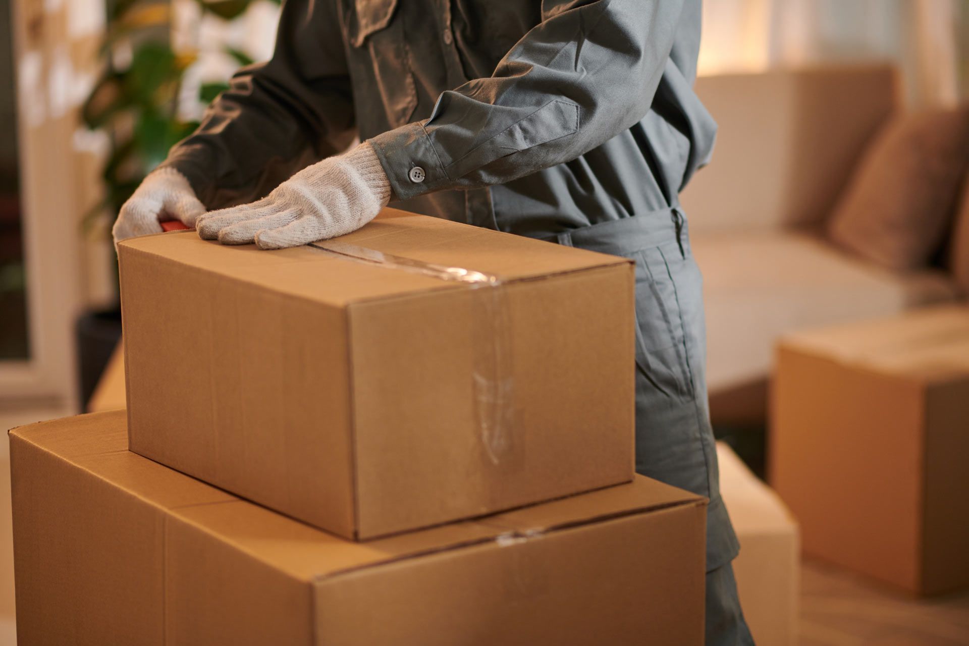 Person in work uniform taping a cardboard box on top of another. Indoors with other boxes and furniture.