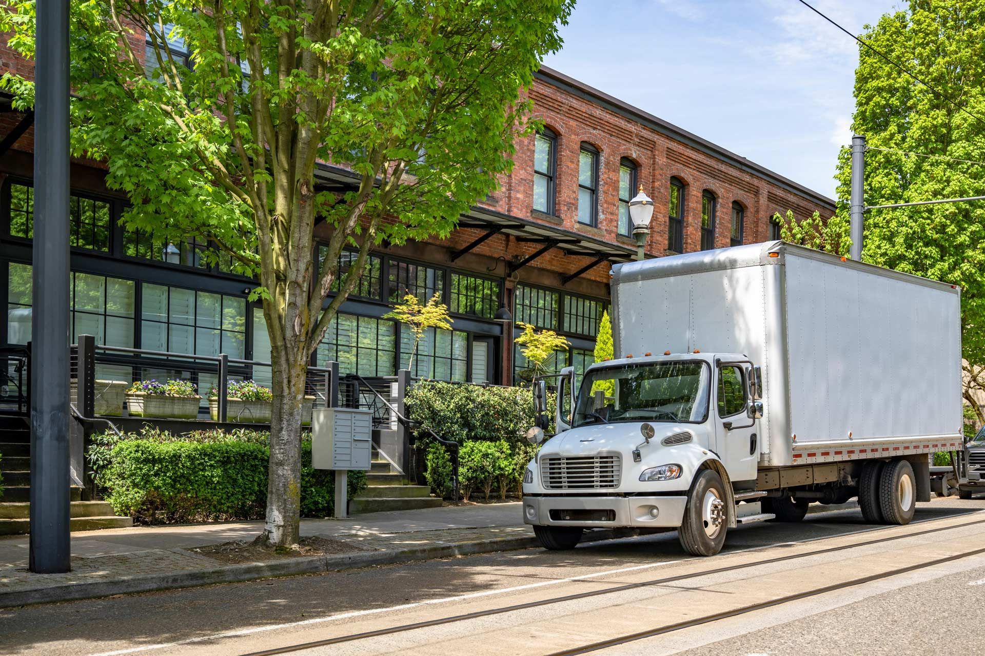 Box truck parked in front of a brick building with glass windows, under green trees, sunny day.