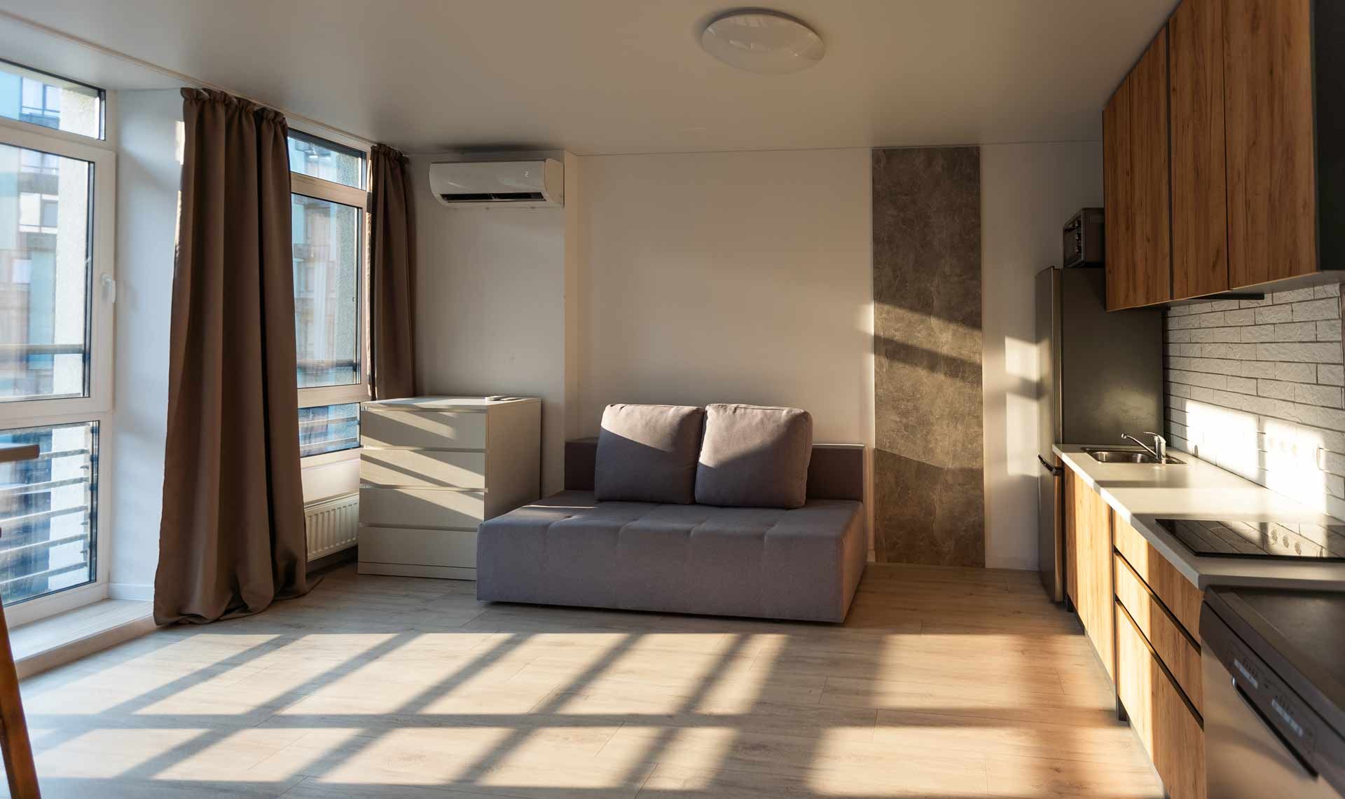 Small apartment interior with sofa, kitchenette, and large window with brown curtains.