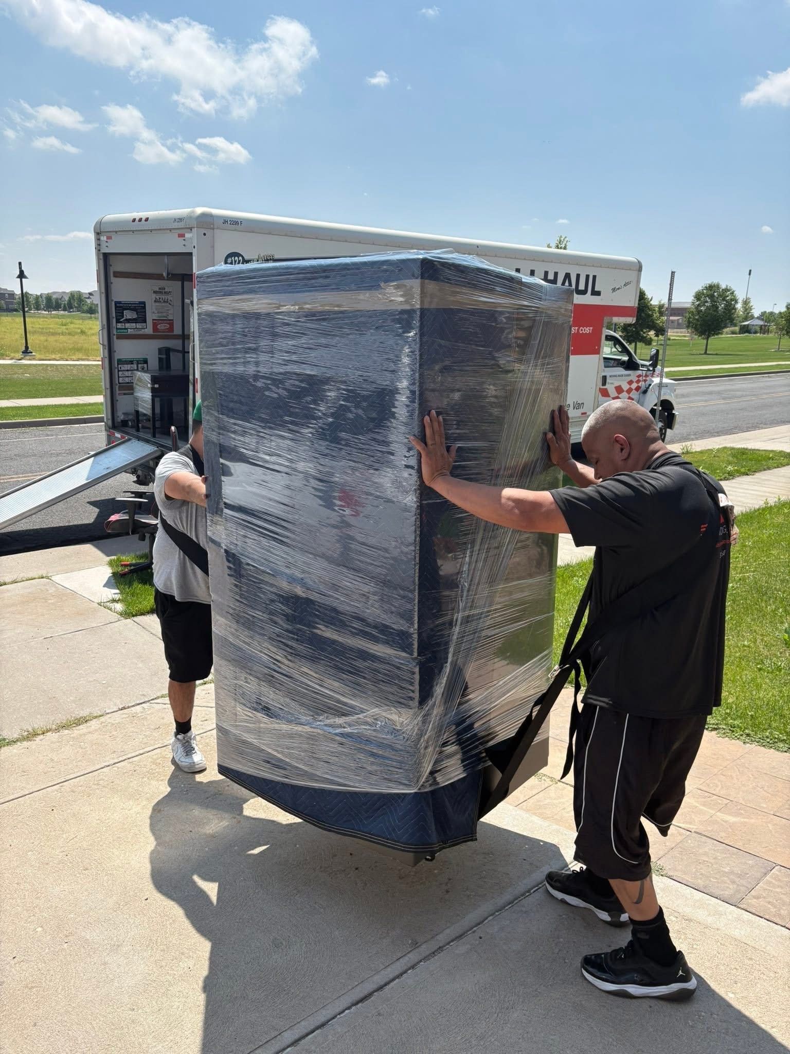 Two people moving a large, wrapped item toward a U-Haul truck on a sunny day.