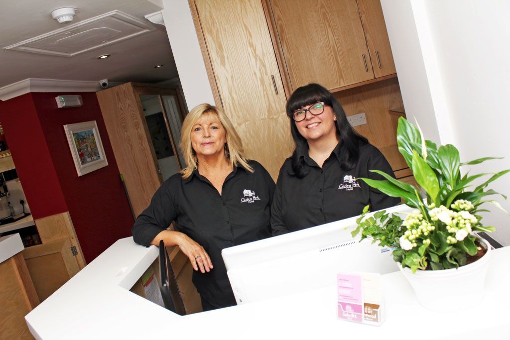 Two women are standing at a reception desk in front of a plant.