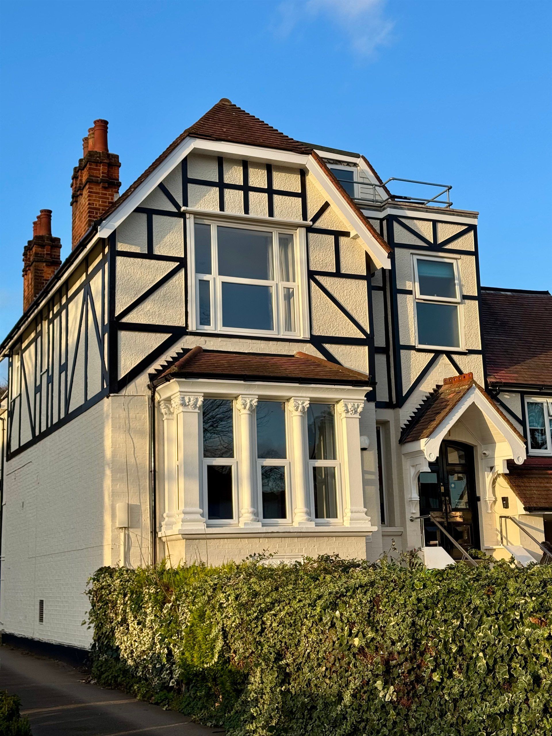 A large white house with a brown roof is surrounded by a hedge.