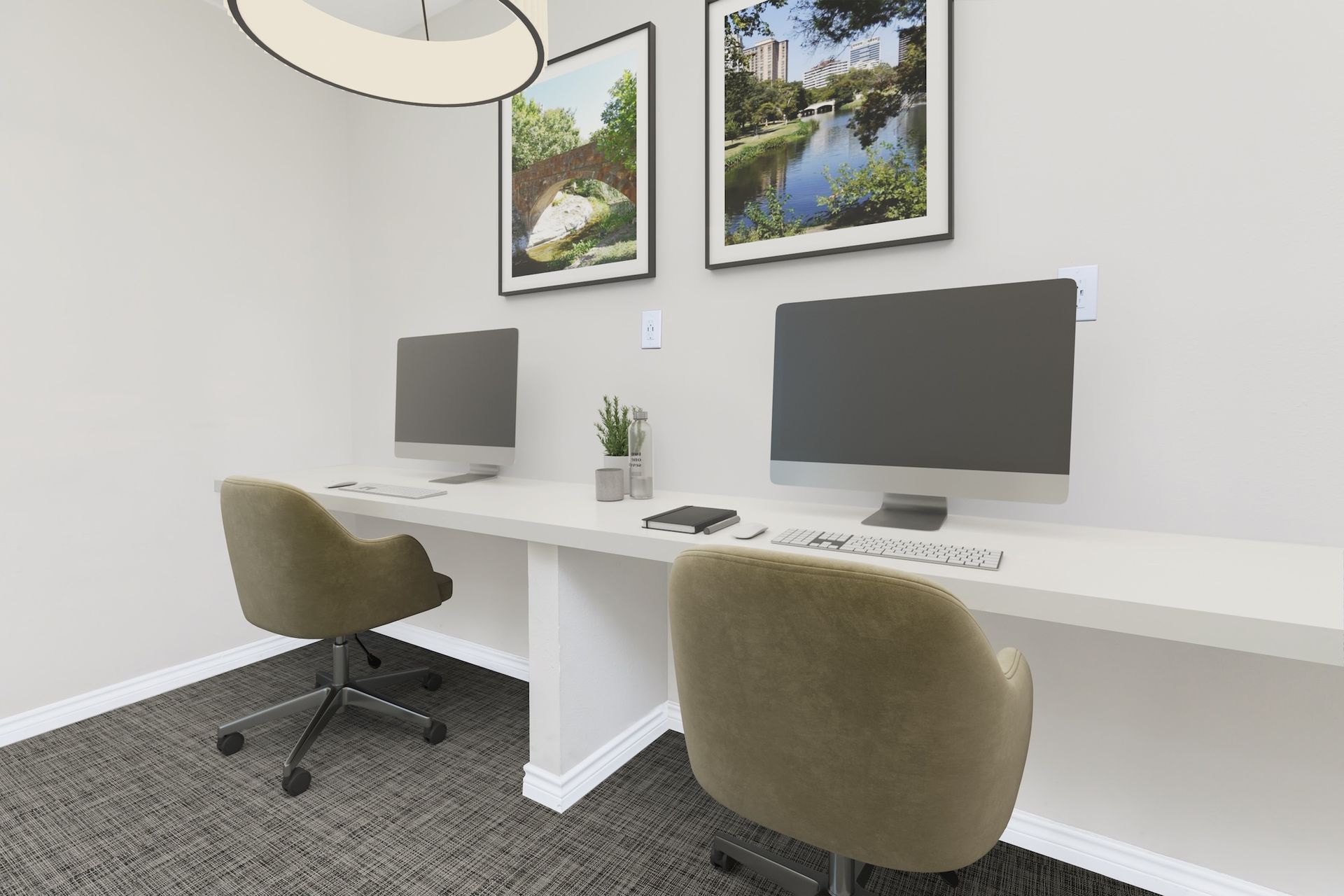 Two computer workstations with monitors, chairs, and decor against a white wall, beneath framed art and a light fixture.