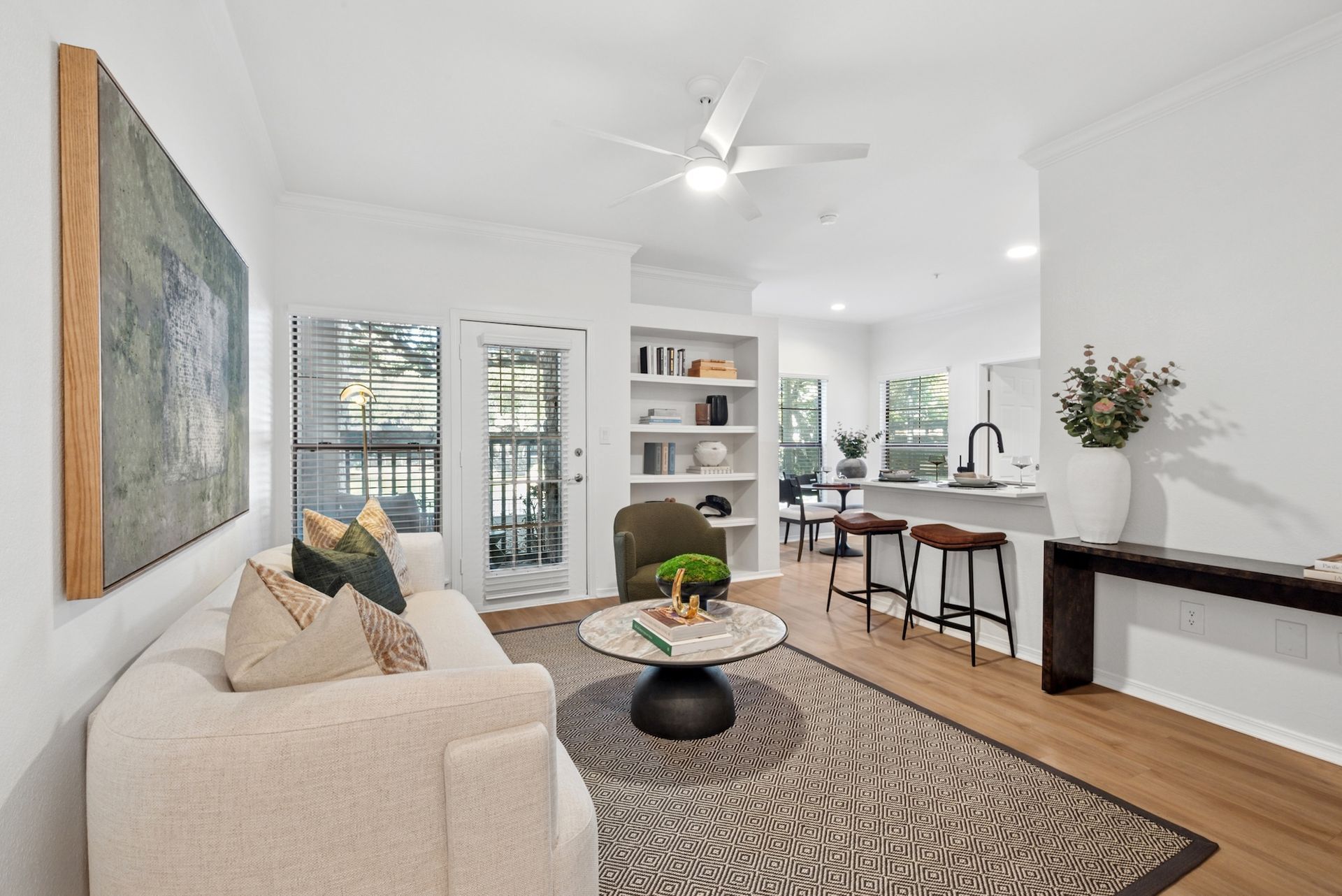 Bright, open-concept living space with neutral decor; white walls, wood flooring, sofa, rug, and a view into the kitchen.