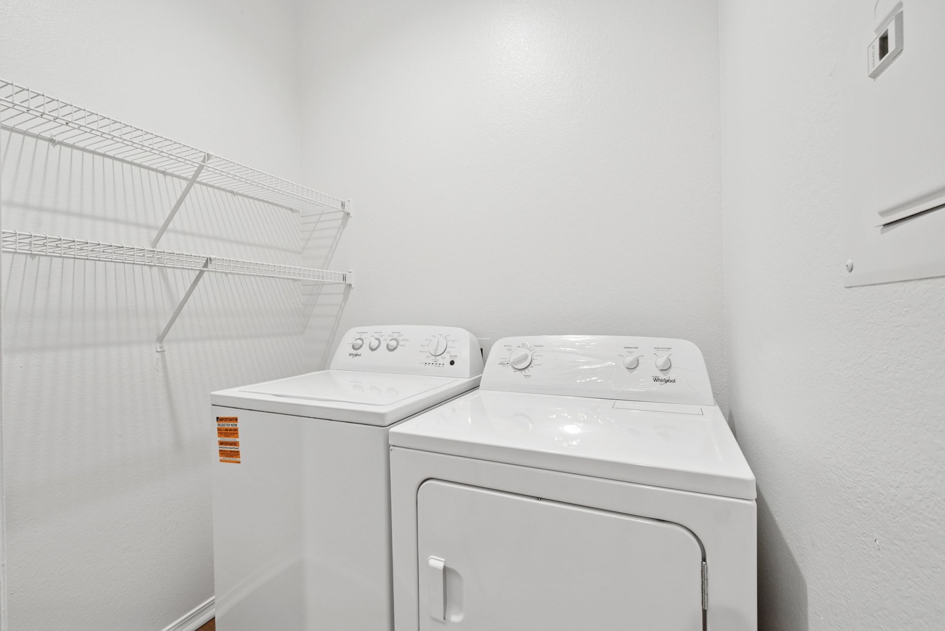 White washer and dryer in a bright laundry room, with wire shelving on the wall.
