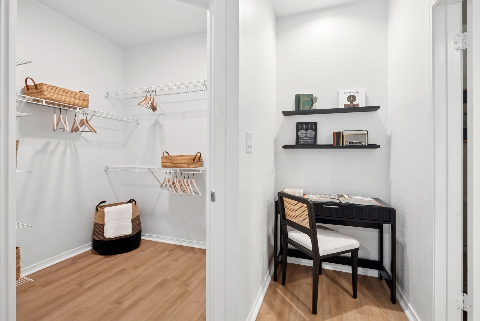 A modern walk-in closet with wire shelving and a small desk. White walls, light wood floors.