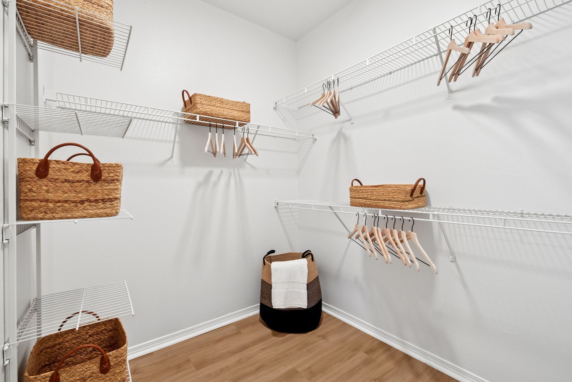 A walk-in closet with wire shelves, baskets, and hangers. The walls are white and the floor is wood.