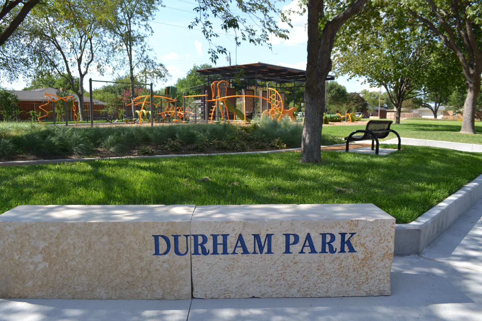 Durham Park sign in front of green grass and playground equipment under a blue sky. A bench is in the middle ground.