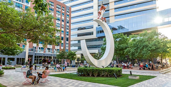 People relax in a plaza with a modern sculpture in the center, surrounded by buildings and trees.